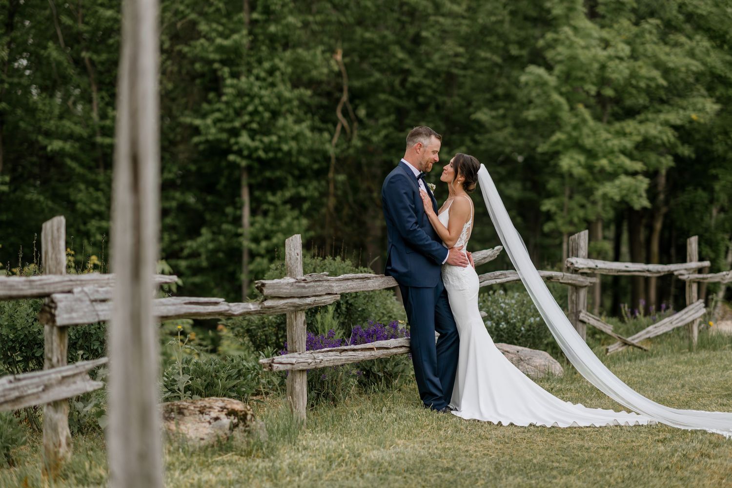 Long white wedding veil trails across rustic wooden fence in outdoor garden setting.