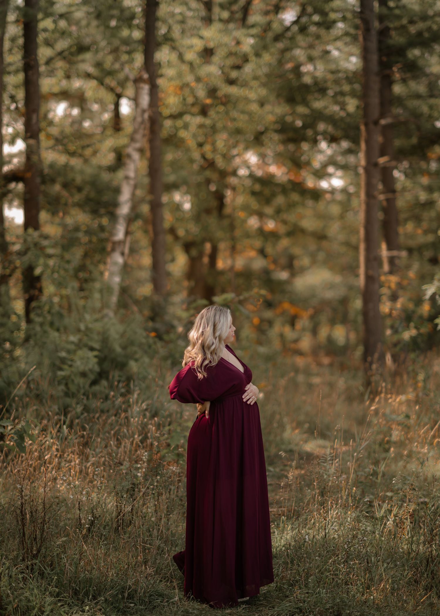 Woman in burgundy dress stands in golden-lit forest during maternity session.