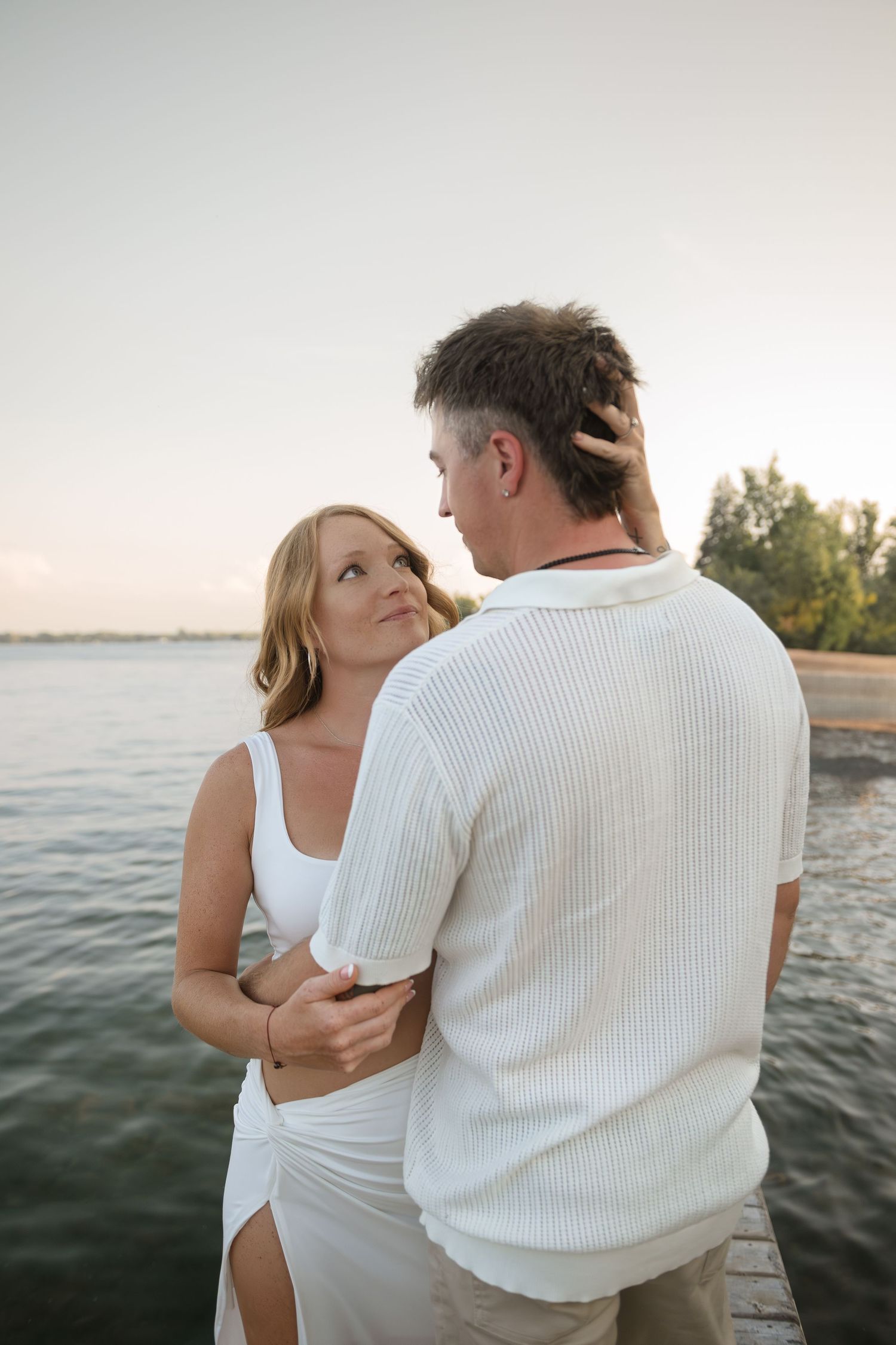 Couple in white outfits shares tender moments by waterfront during sunset.