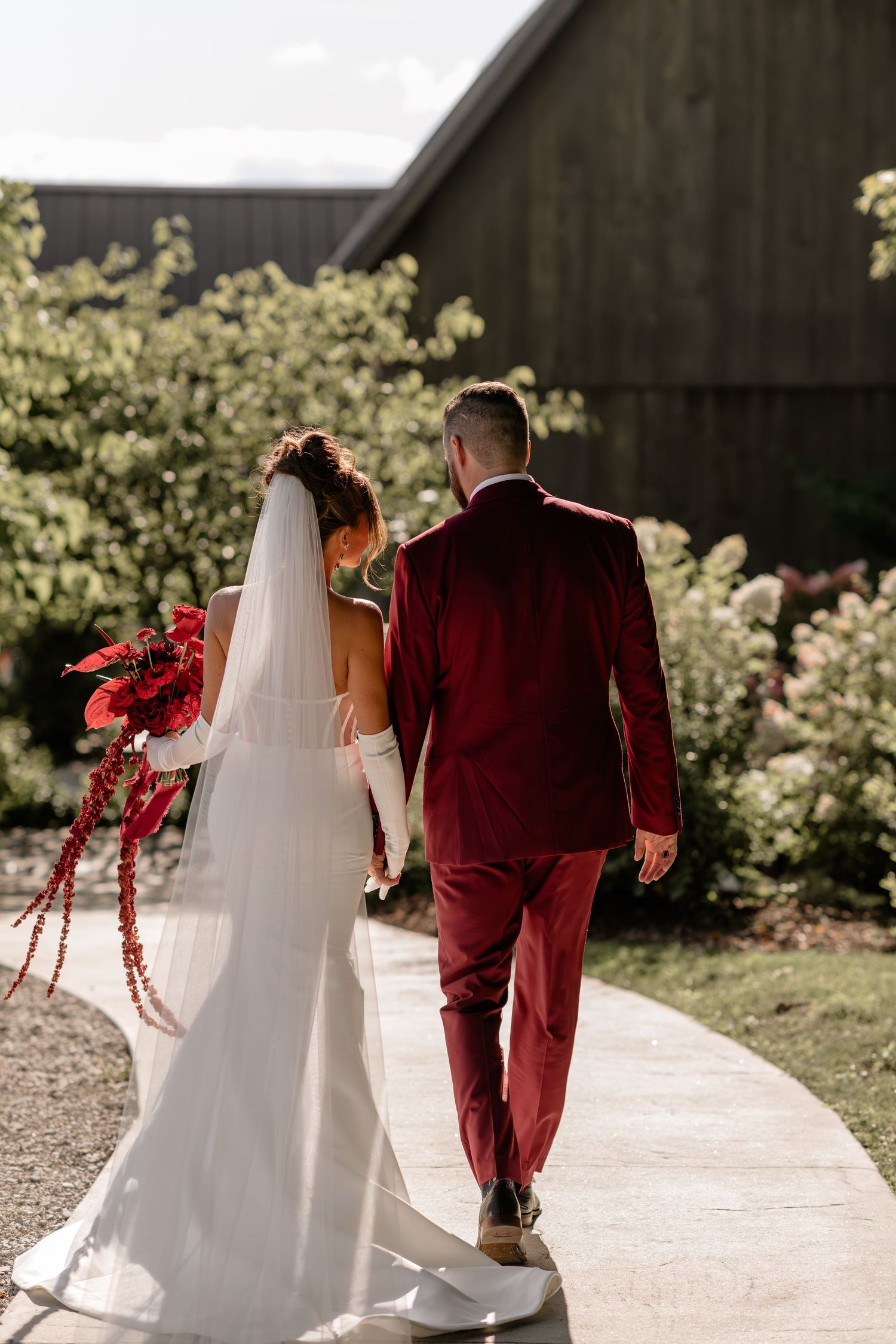 A bride in white and groom in burgundy walk away holding hands on a stone path.