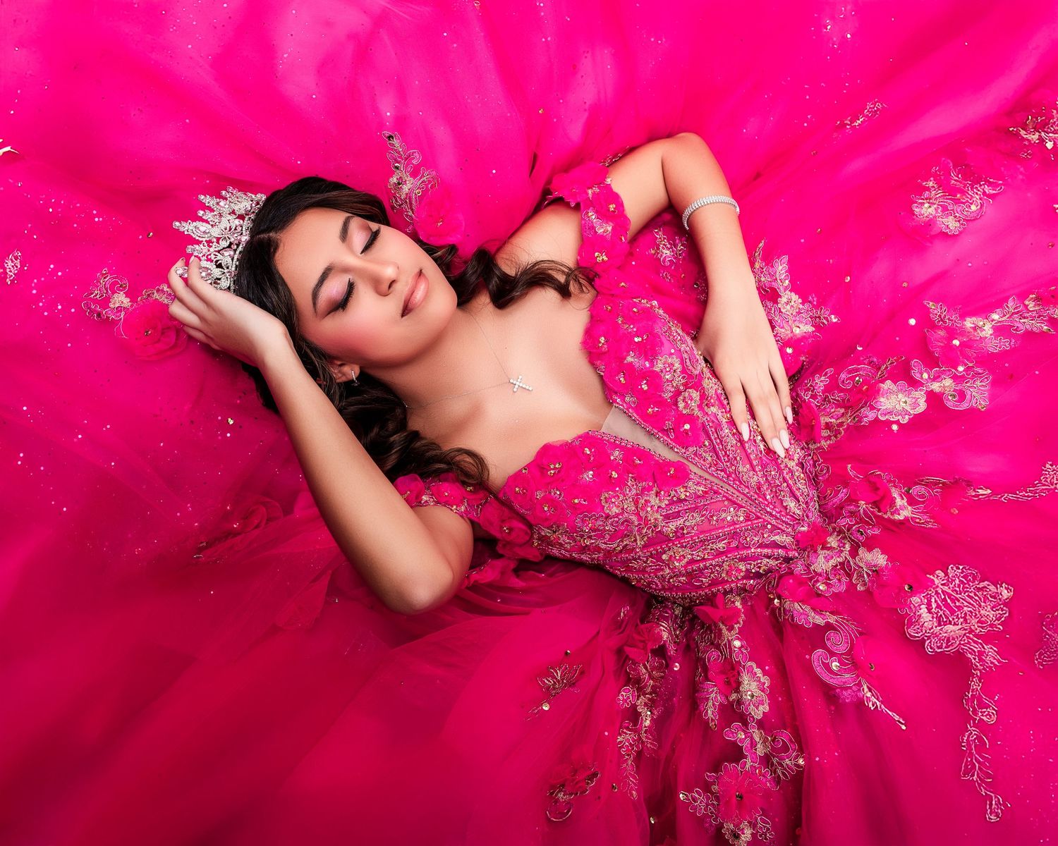 Young woman in a pink quinceañera ball gown with floral embroidery and tiara, lying gracefully on her dress. Sweet 16 photography