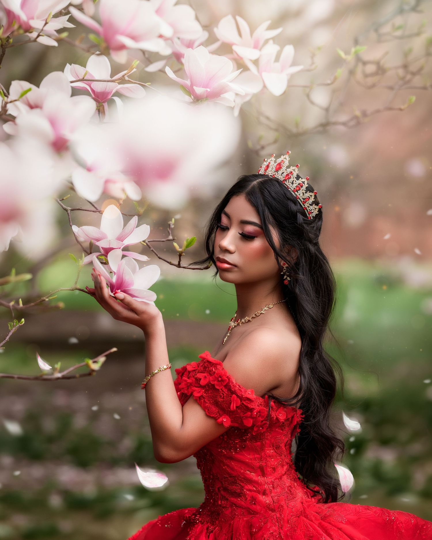 Young woman in red off-shoulder gown and crown holds magnolia blooms among flowering spring trees.