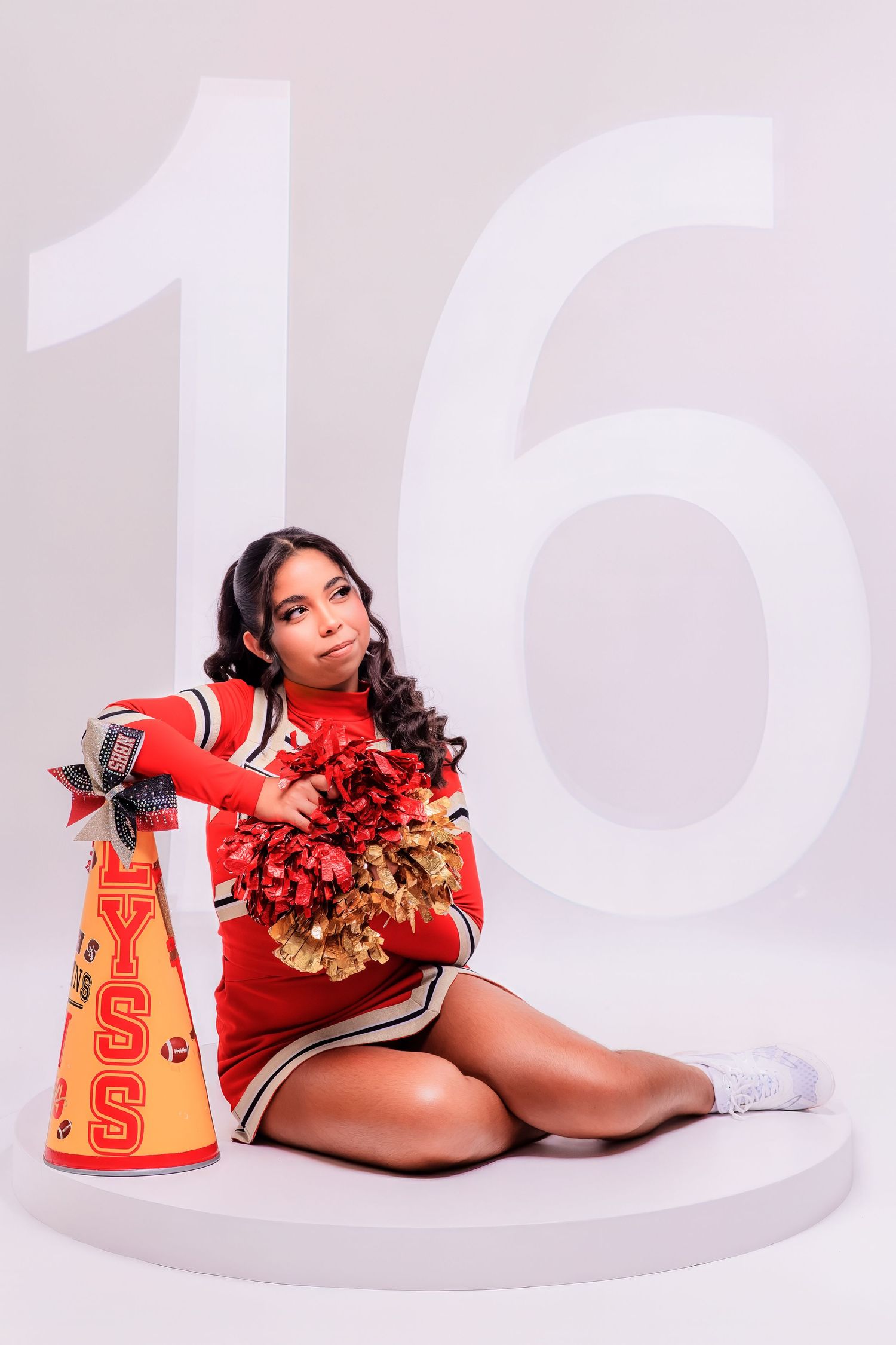 A cheerleader in a red uniform sits on a white platform holding red and gold pom-poms next to a megaphone with the number 16 behind her.