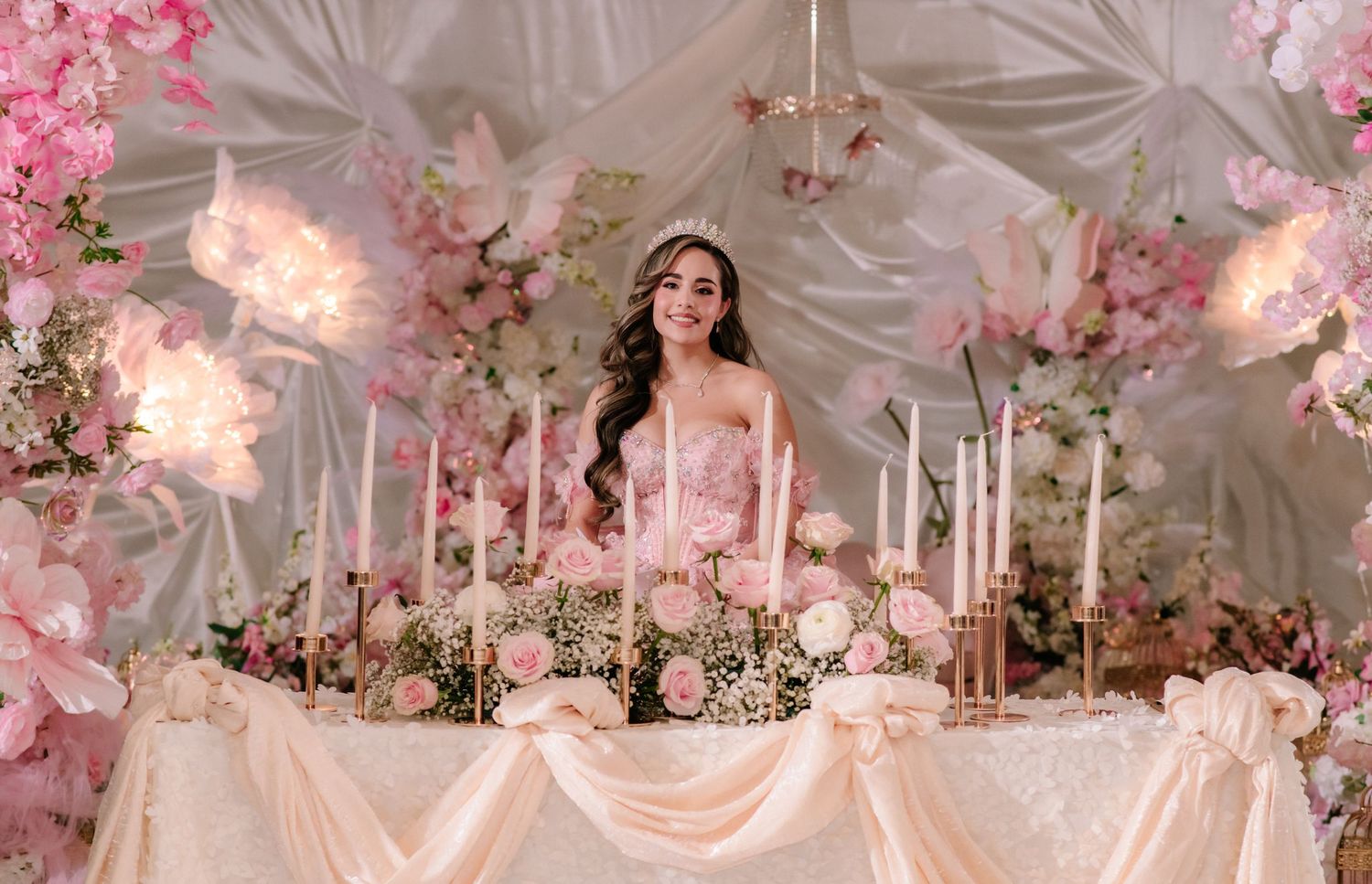 Young woman in pink gown wearing tiara sits at elegant floral reception table with candles and pink roses.