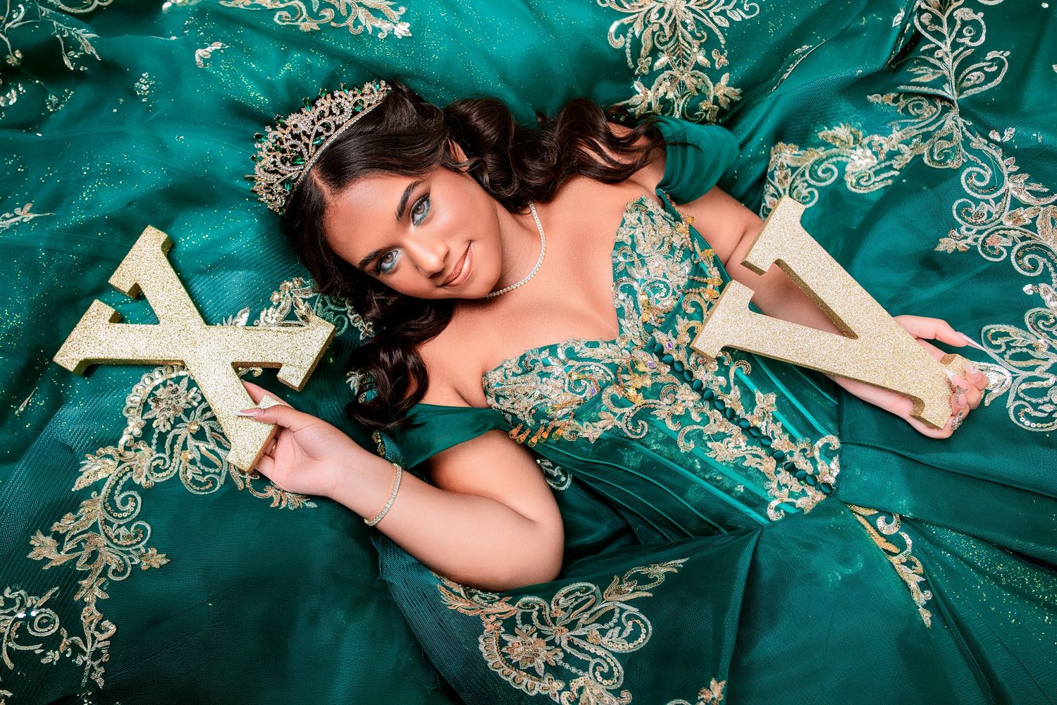 Young woman in emerald green quinceañera gown with gold embroidery and tiara holds decorative letters for a portrait.