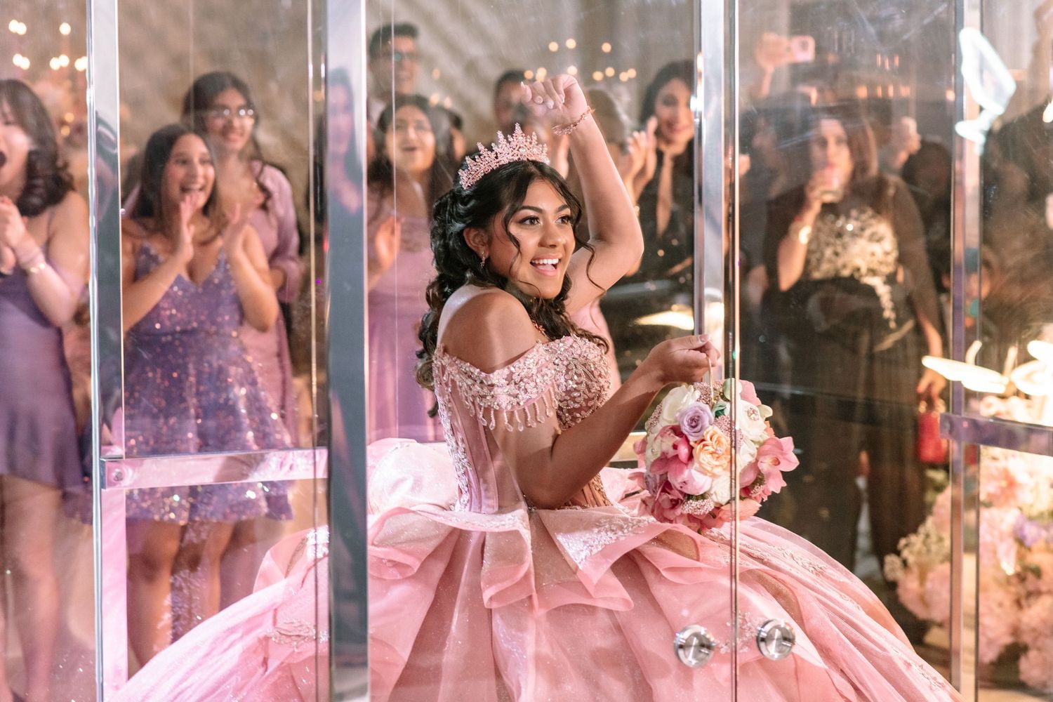 Young girl in pink quinceañera ball gown wearing tiara holds bouquet surrounded by celebrating guests.