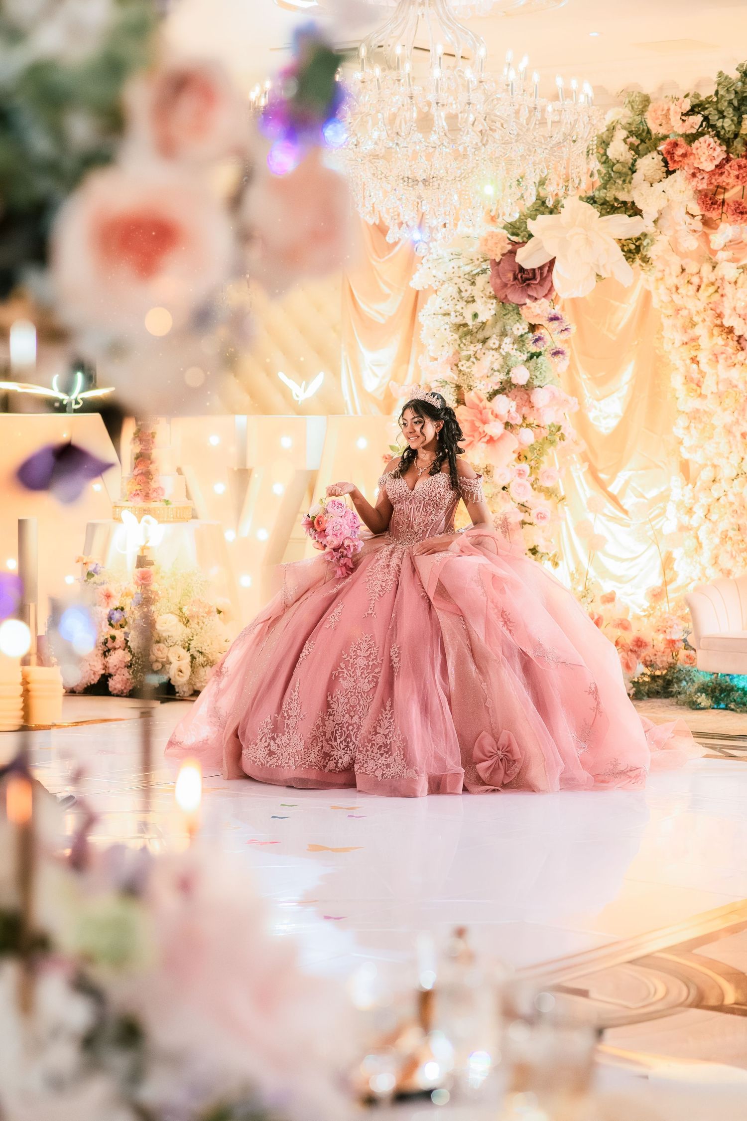 Quinceañera in pink lace ball gown poses on dance floor with elegant floral chandelier backdrop.