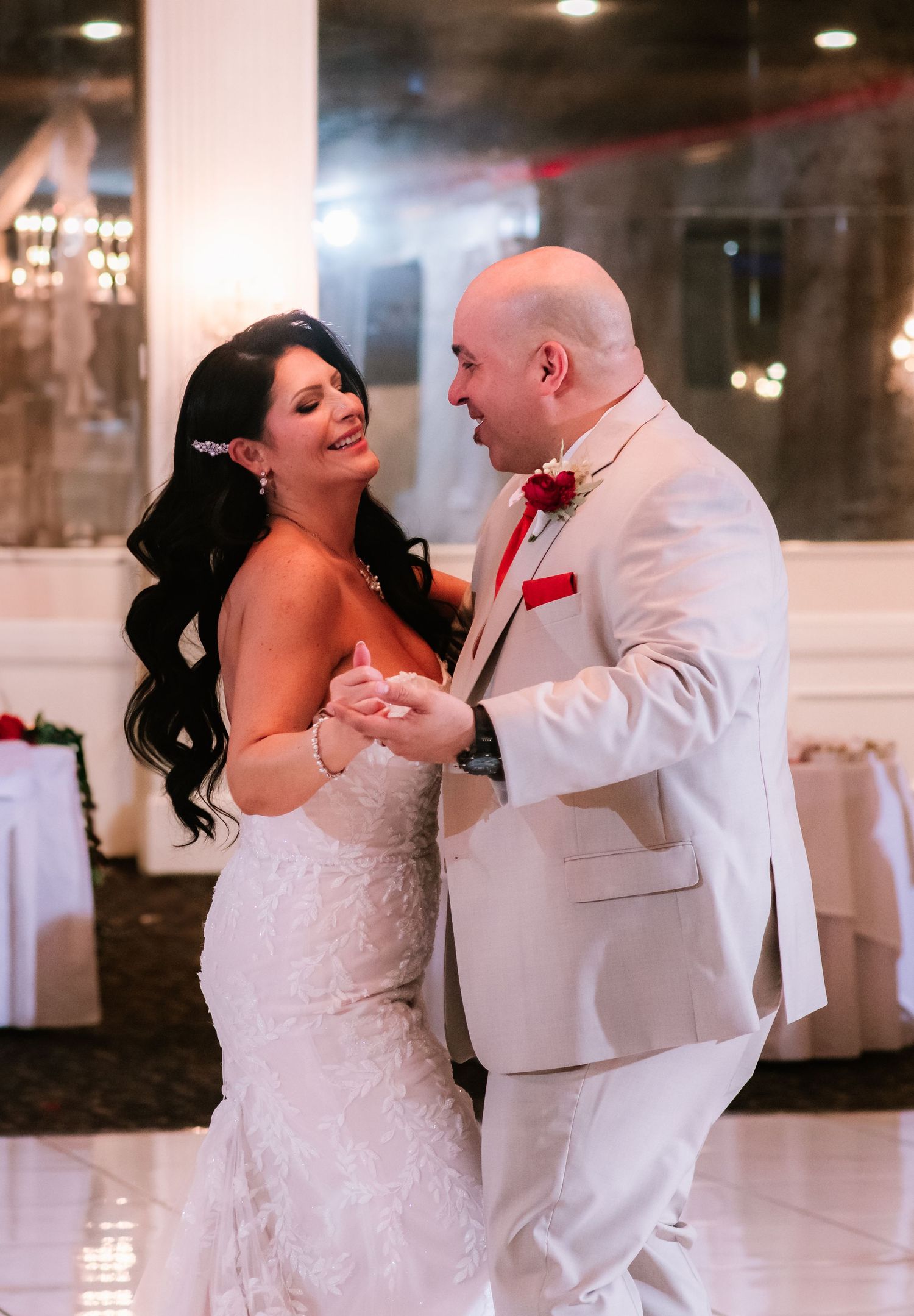 Bride and groom share their first dance at wedding reception, wearing white lace gown and cream tuxedo with red boutonniere.