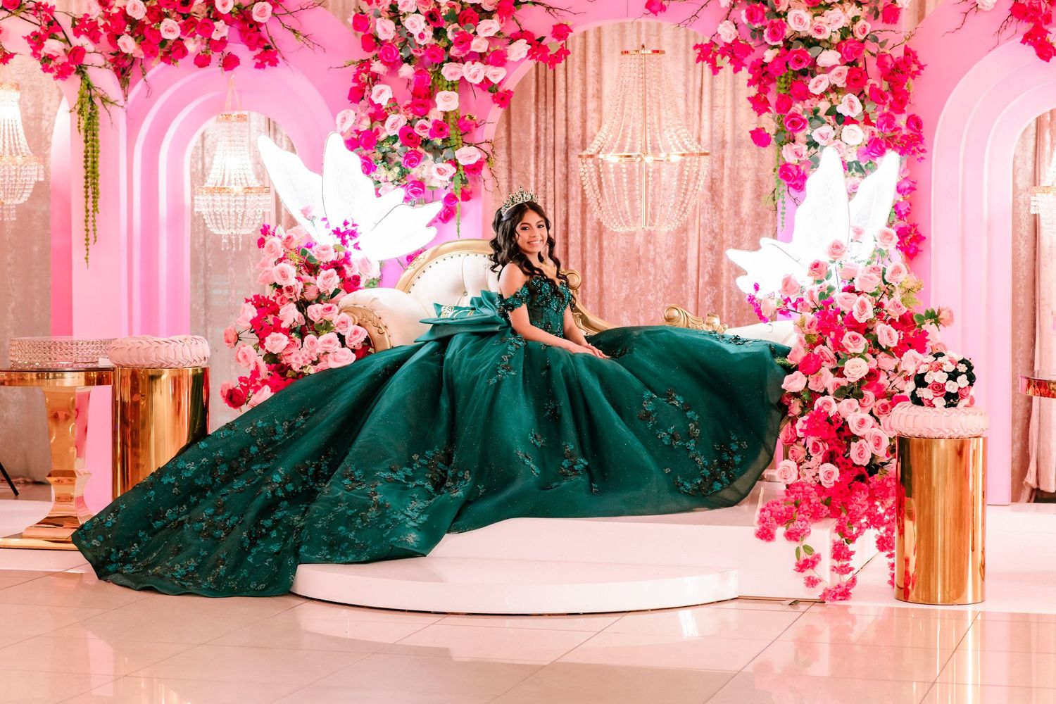 Young woman in emerald green quinceañera ball gown poses on white stage surrounded by pink floral decorations.