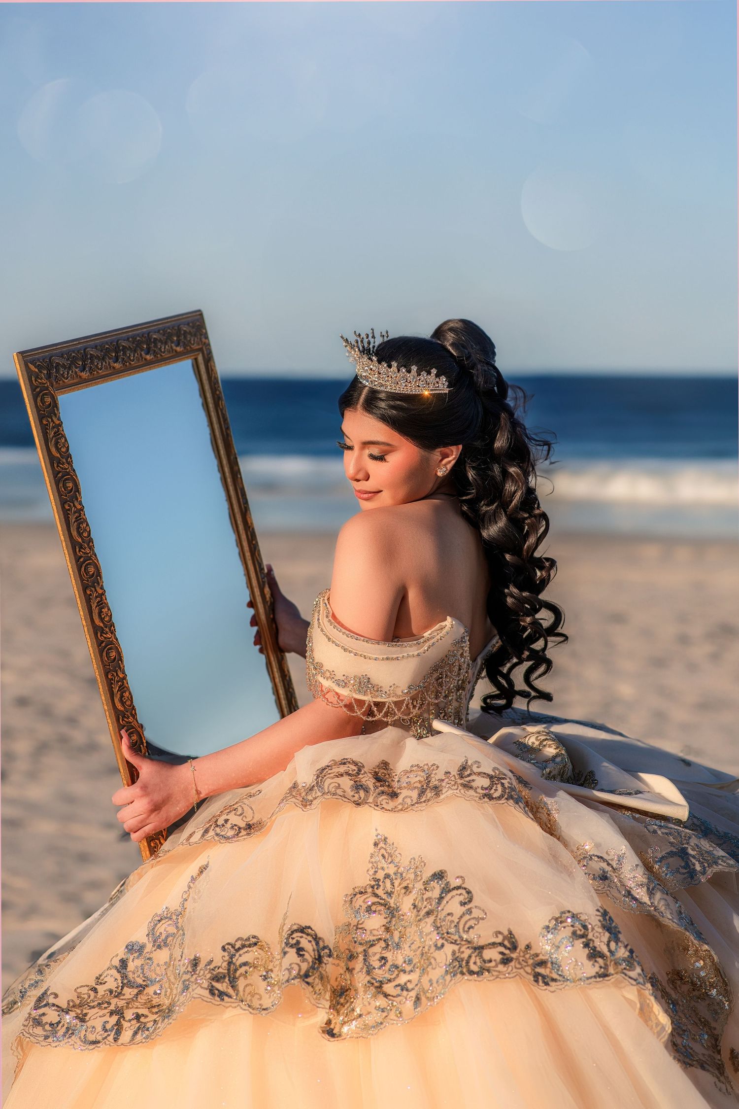 A young woman in a gold quinceañera gown and crown holds an ornate mirror on a sunny beach.