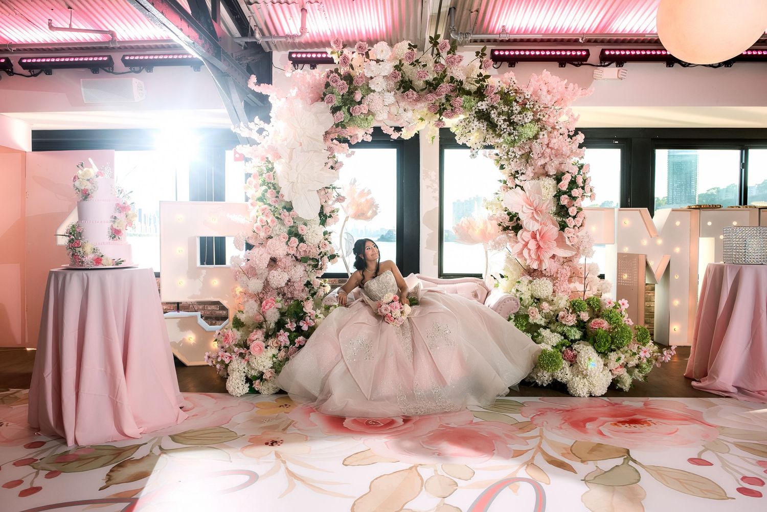 Young woman in a pink ball gown poses under a floral arch at an elegant pink-themed quinceañera celebration.