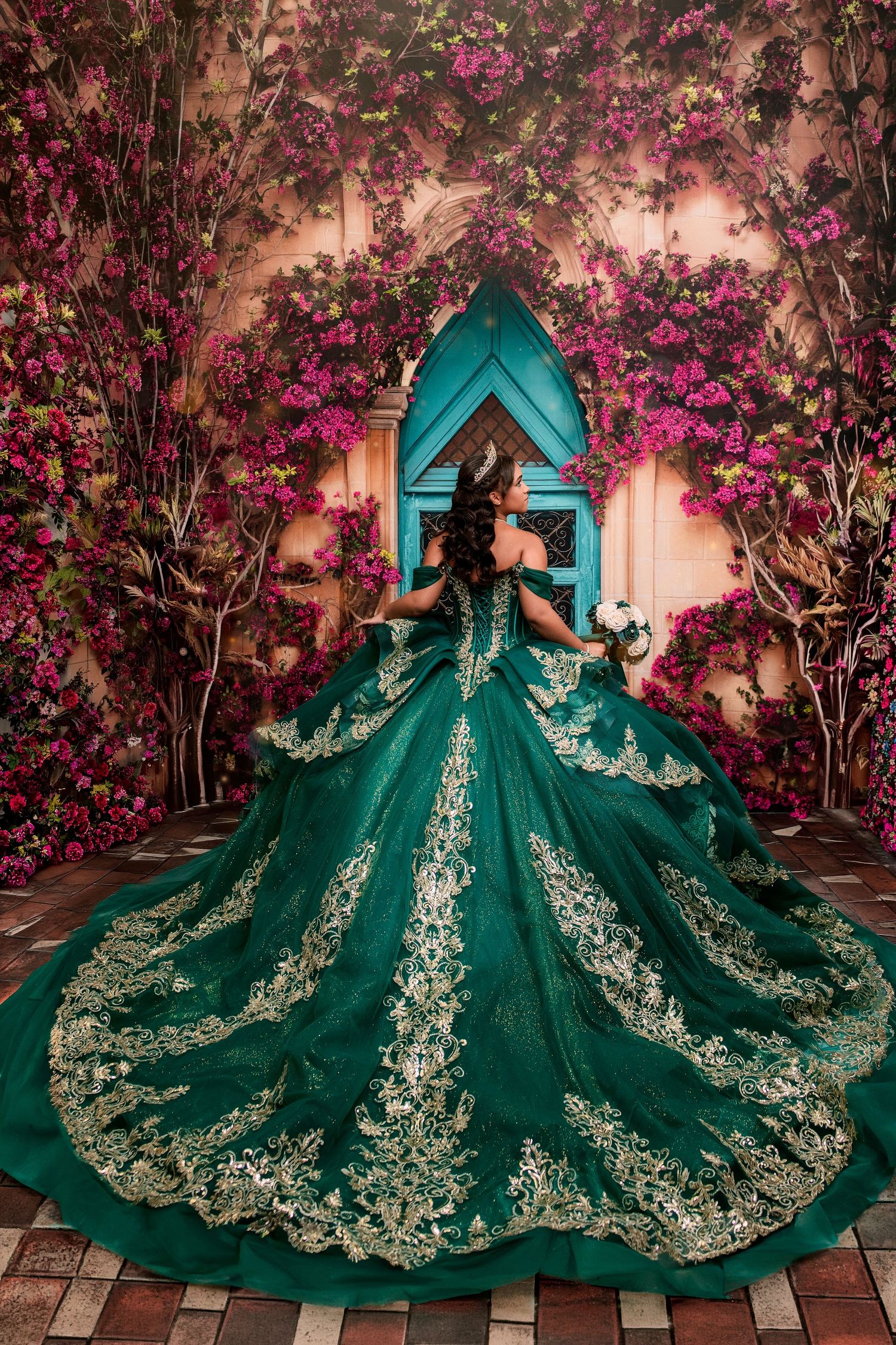 Woman in emerald green quinceañera ball gown with gold embroidery stands before floral arch with pink bougainvillea blooms.