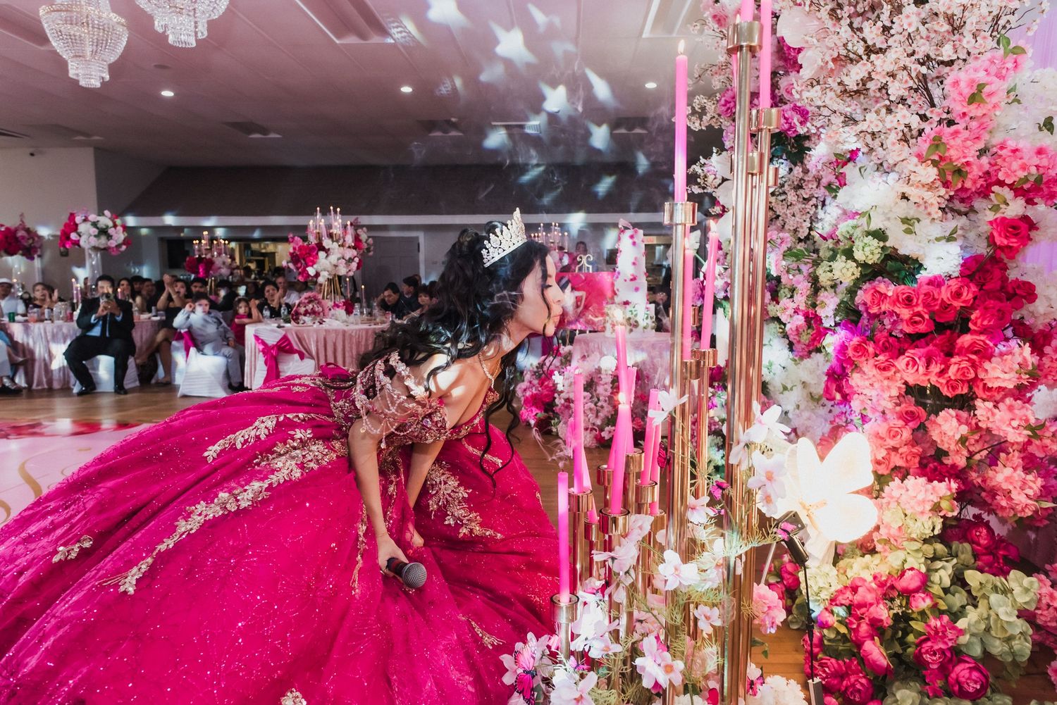 A girl in a stunning pink quinceañera gown and tiara leans toward an elaborate floral arrangement at her celebration.