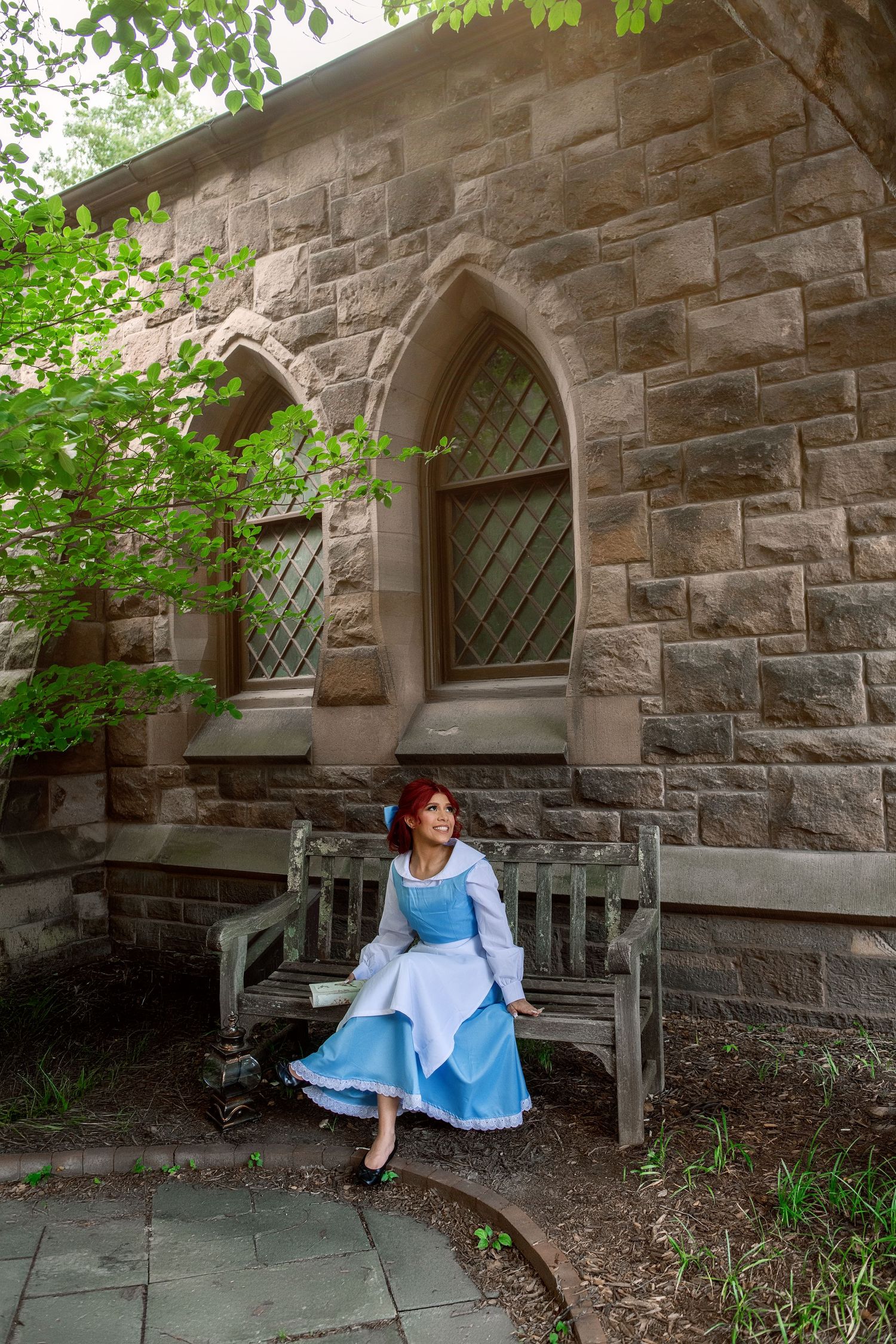 Woman in blue Ariel costume sits on wooden bench beside stone castle wall with Gothic arch windows.