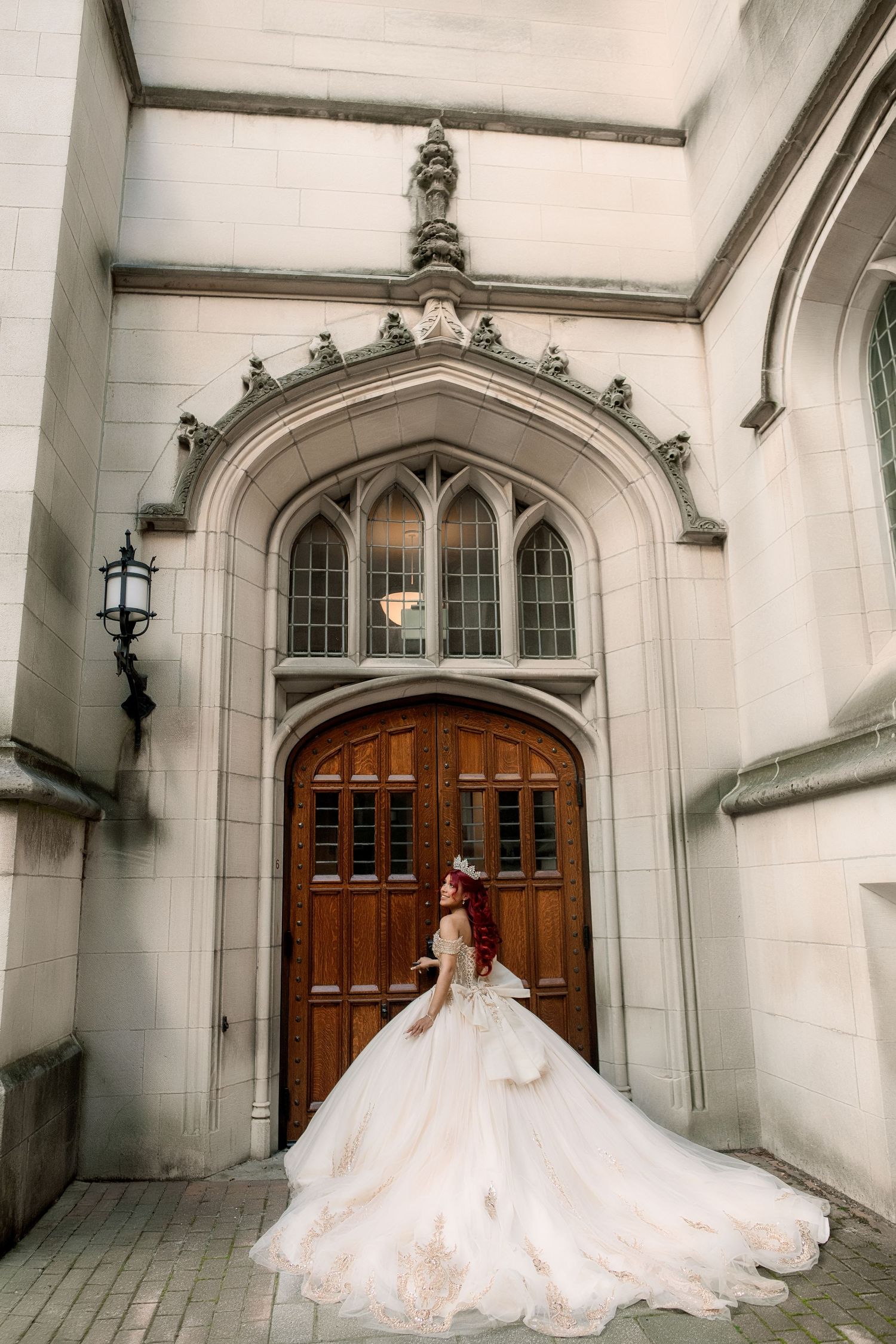 A bride in a flowing white ballgown stands before an ornate Gothic stone archway with wooden doors.