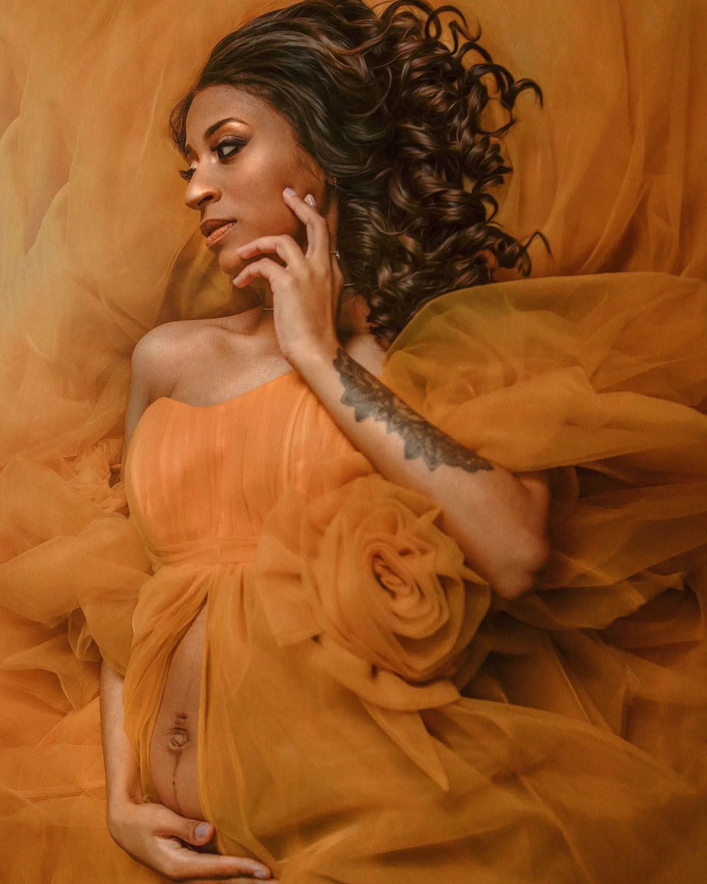 Pregnant woman in elegant orange tulle gown posing gracefully with curly hair and floral detail.