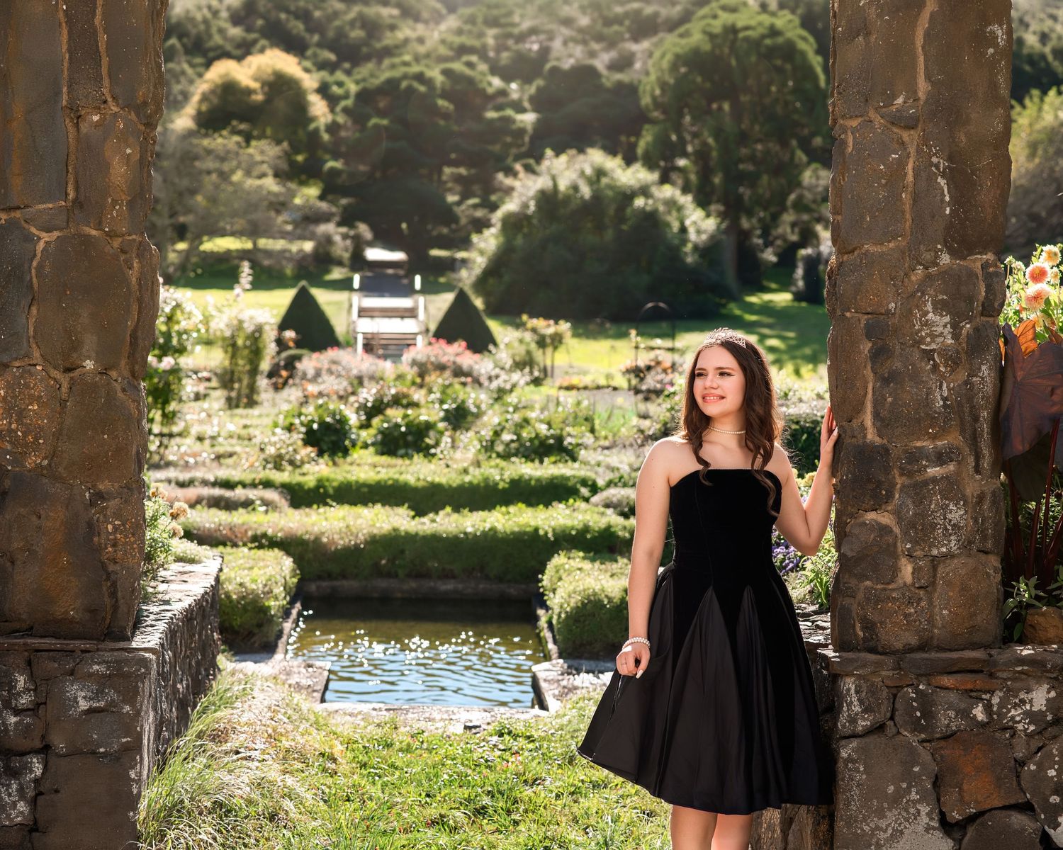 Young woman in black dress posing between stone columns in a lush formal garden.