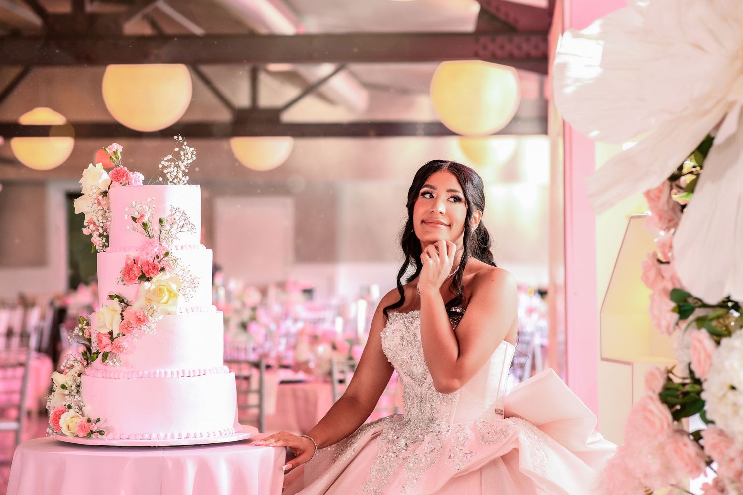 Young woman in a beaded ball gown gazes dreamily beside a floral white tiered cake at an elegant pink quinceañera.