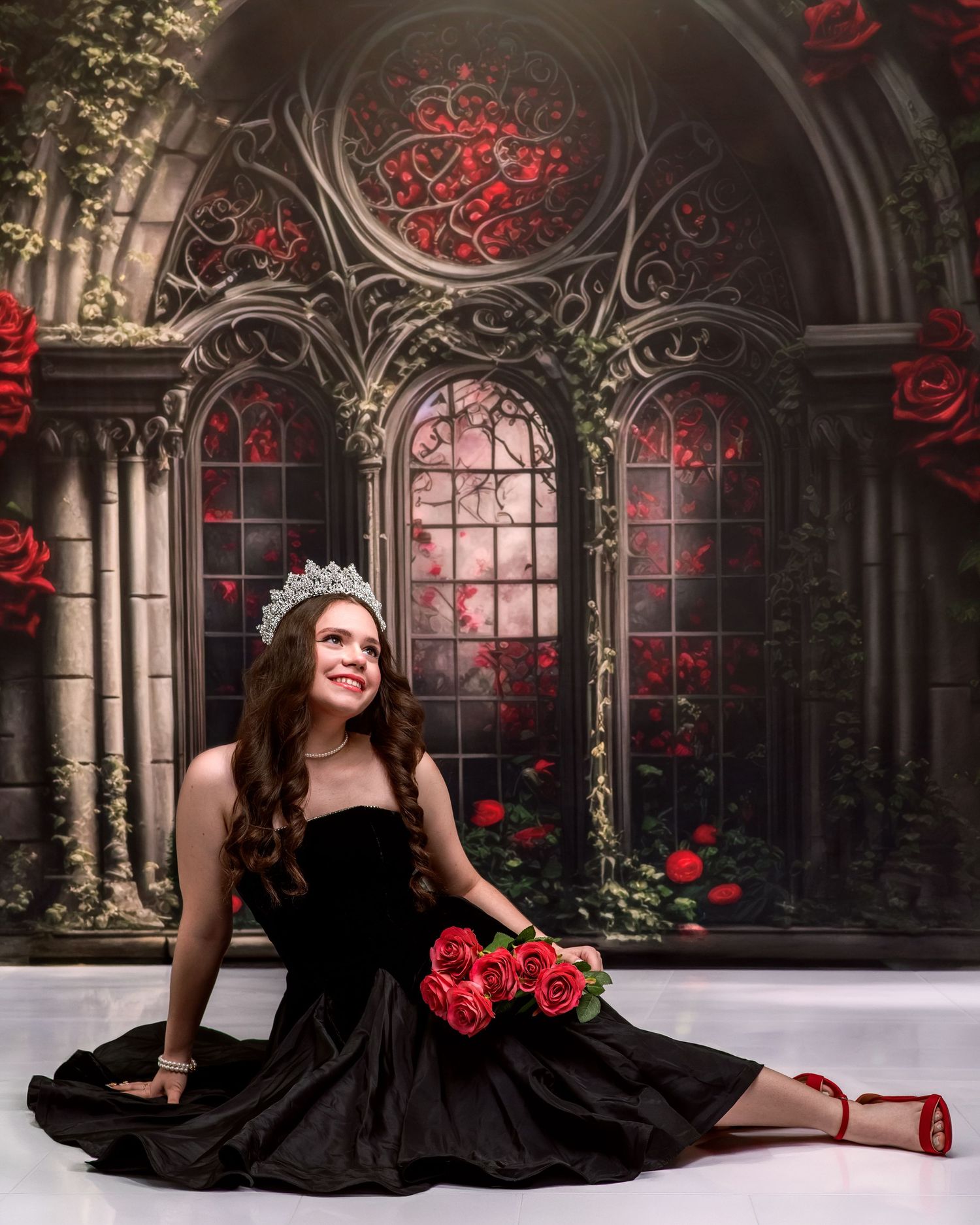 Woman in black gown with tiara holding red roses before gothic rose-adorned backdrop.
