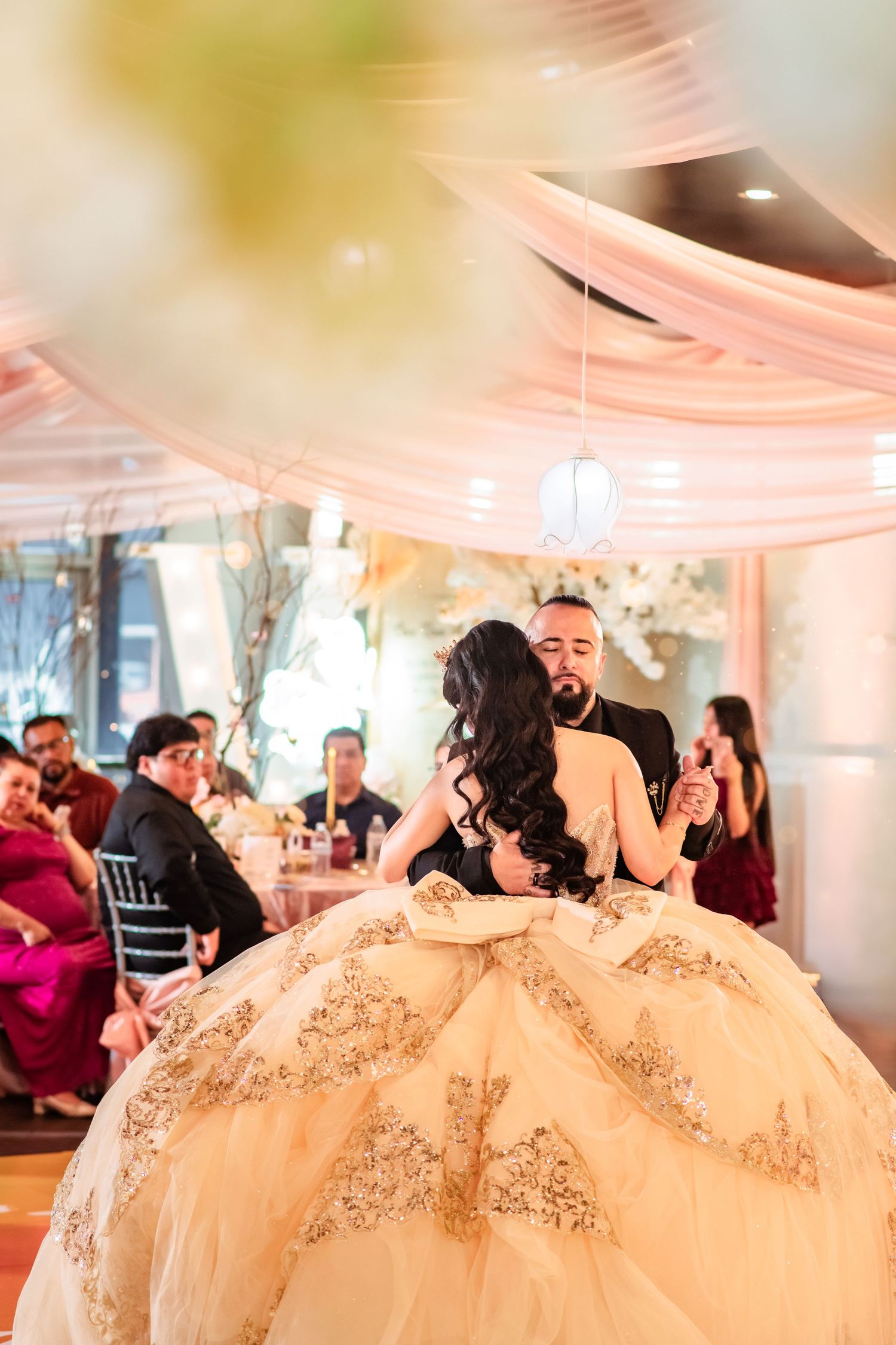 A couple shares their first dance at a wedding reception, the bride wearing an elaborate gold lace ballgown.