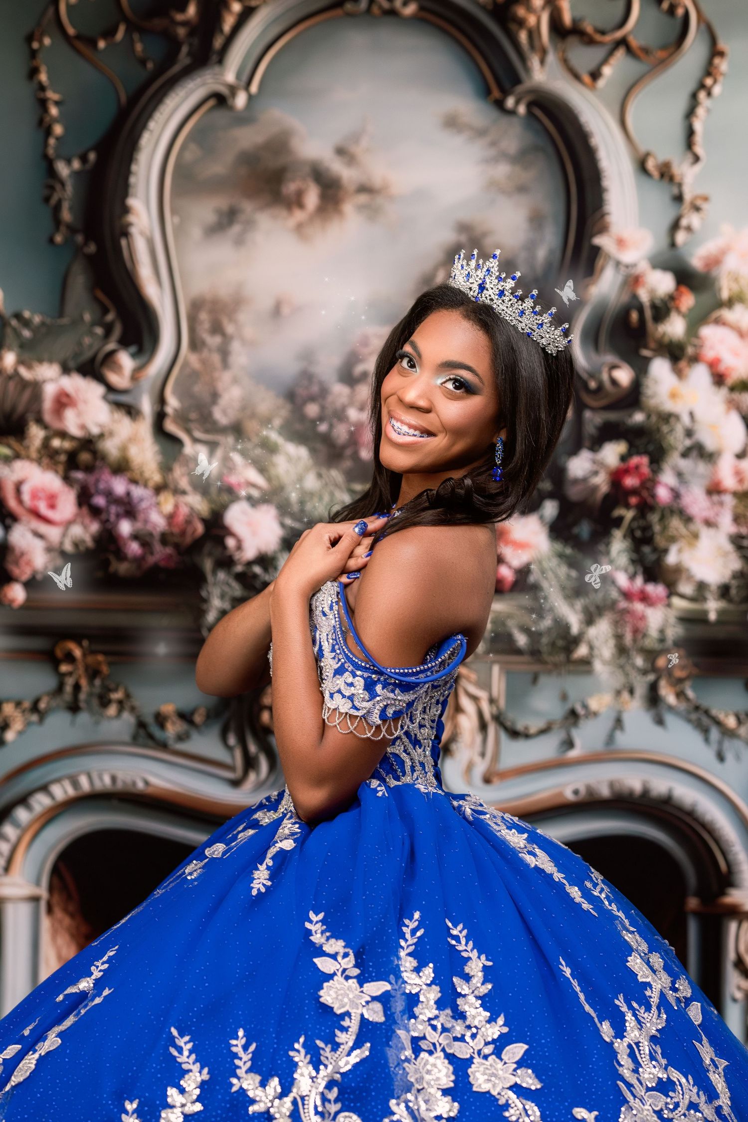 Young woman in royal blue quinceañera gown with silver embroidery and crown poses before ornate floral backdrop.