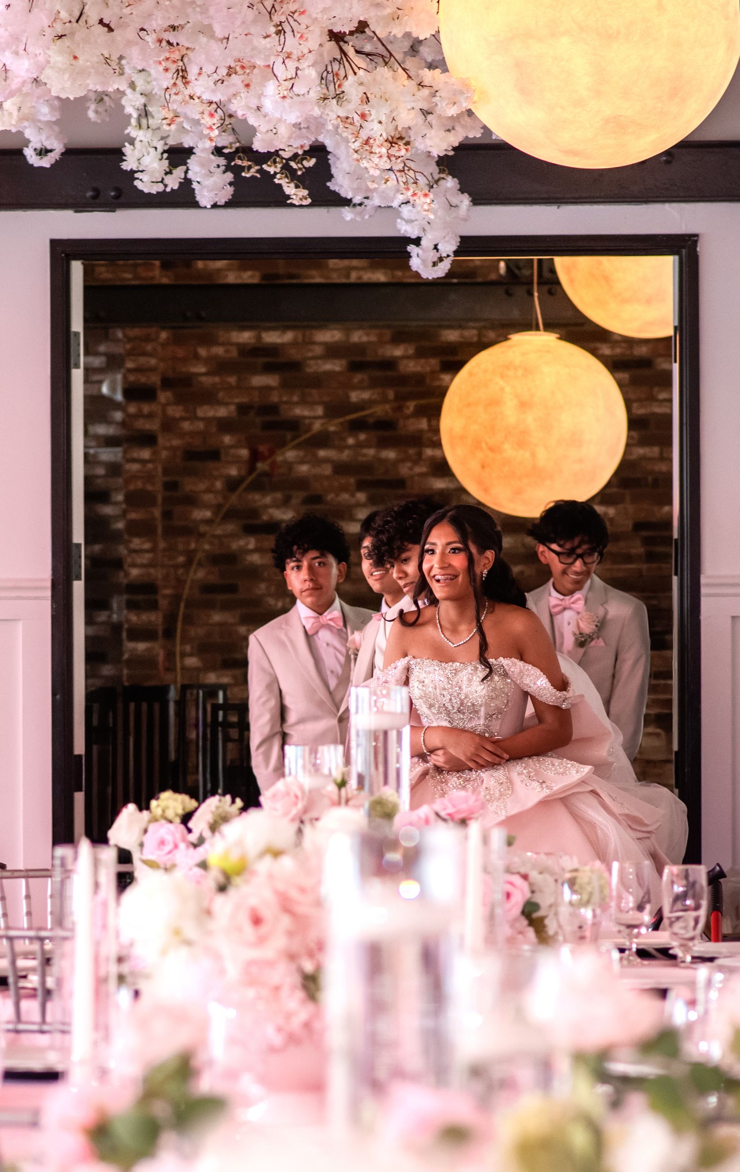 A bride in a pink ball gown smiles with groomsmen in a floral-decorated wedding venue with glowing moon lanterns.