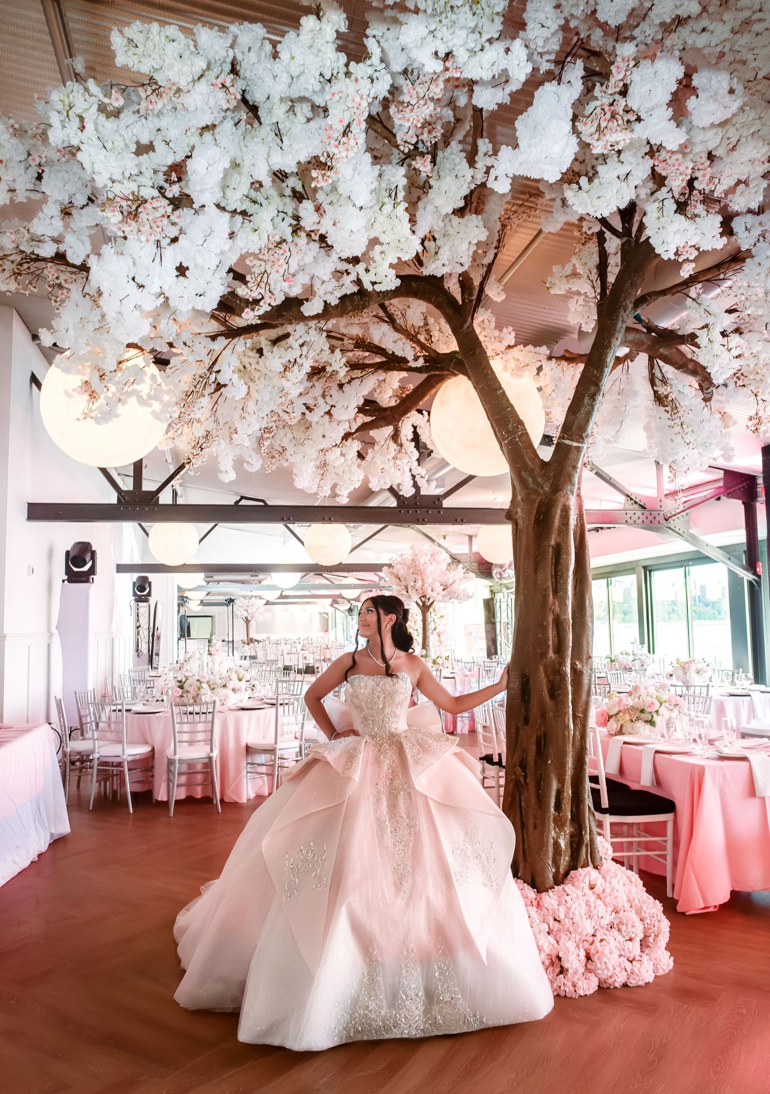 Young woman in a pink ball gown quinceañera dress poses under a white cherry blossom tree in an elegant pink reception hall.