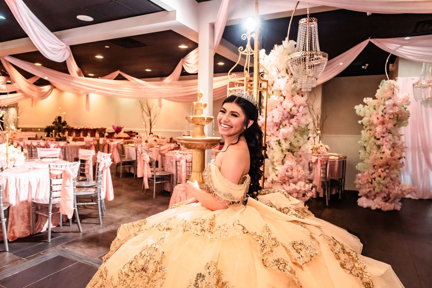 Young woman in elegant gold quinceañera gown poses in decorated pink and white event venue with chandelier.