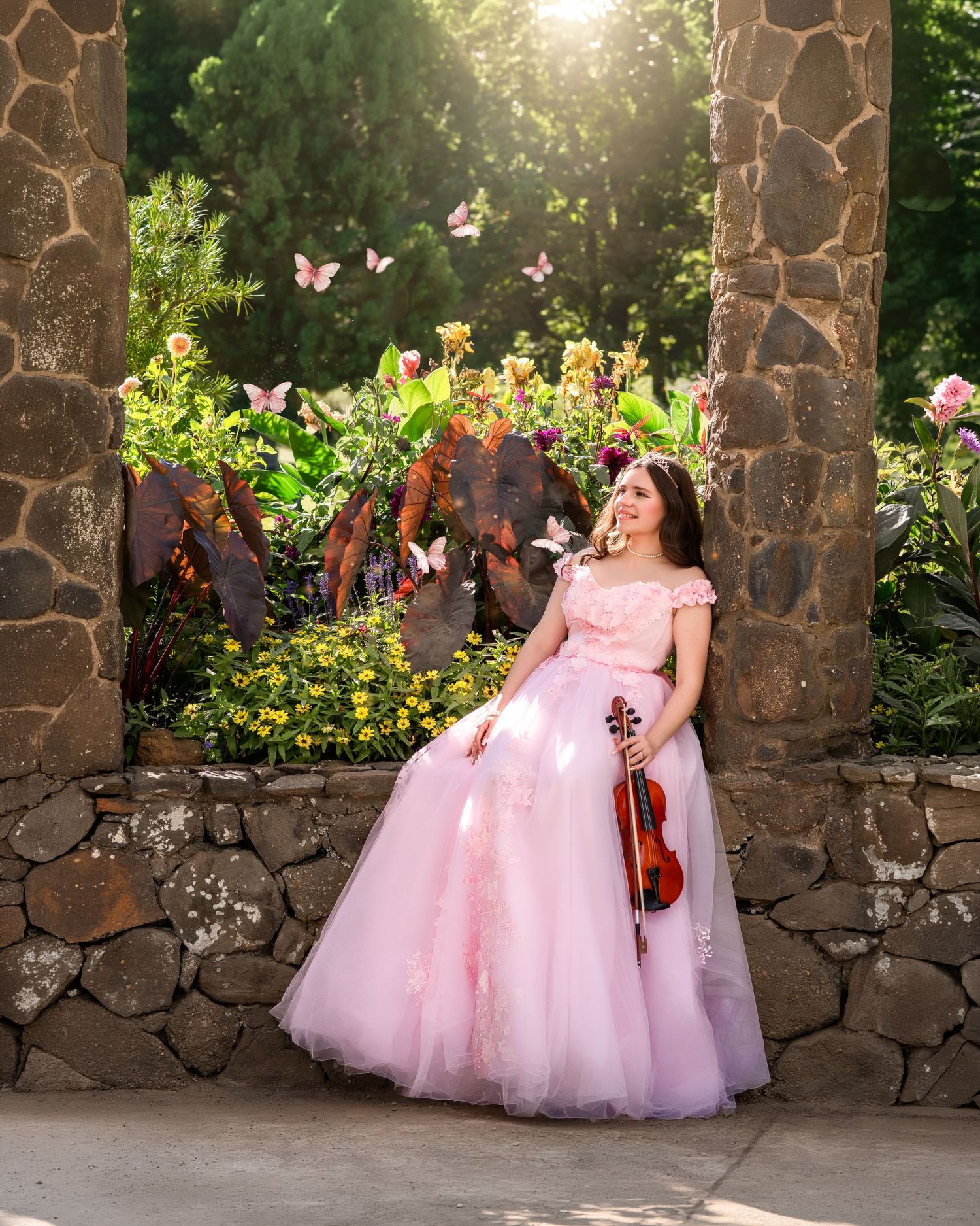 Young woman in pink ball gown holding violin poses in sunlit stone garden with butterflies and colorful flowers.