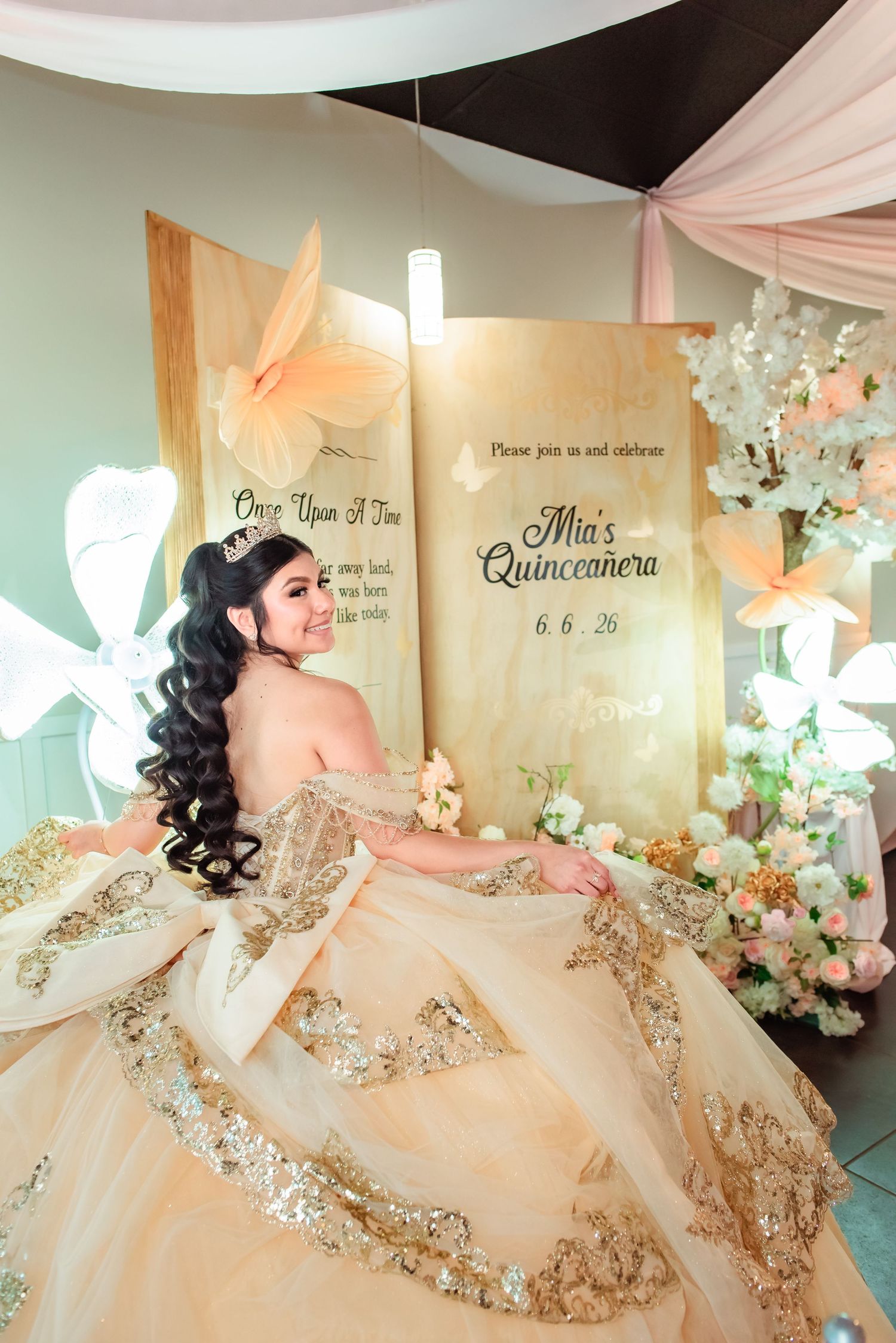 Young woman in gold quinceañera gown poses before decorative backdrop celebrating Mia's Quinceañera on 6.6.26.