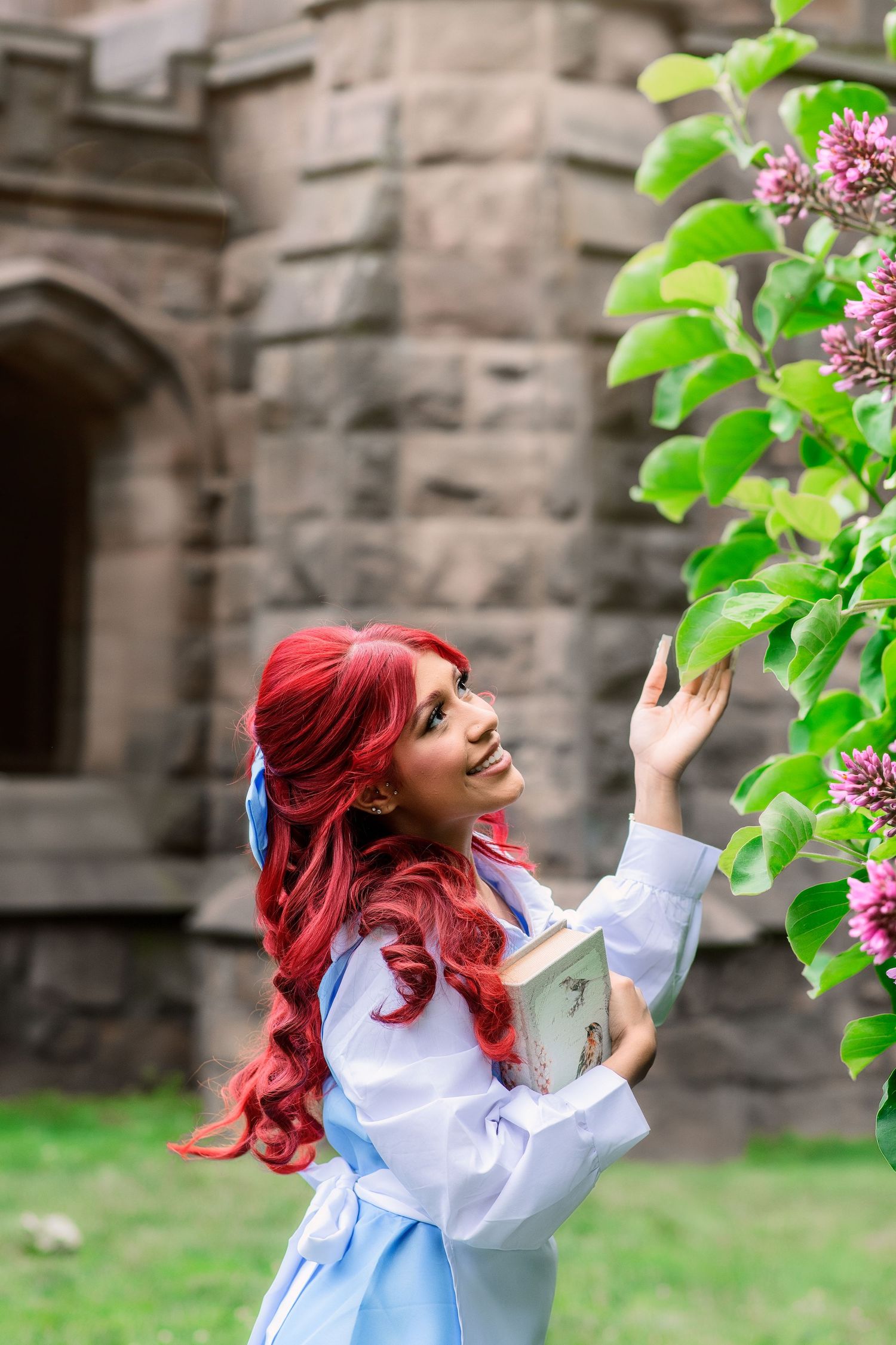 Woman in red wig and blue dress holds book near blooming lilac bush by stone castle backdrop.