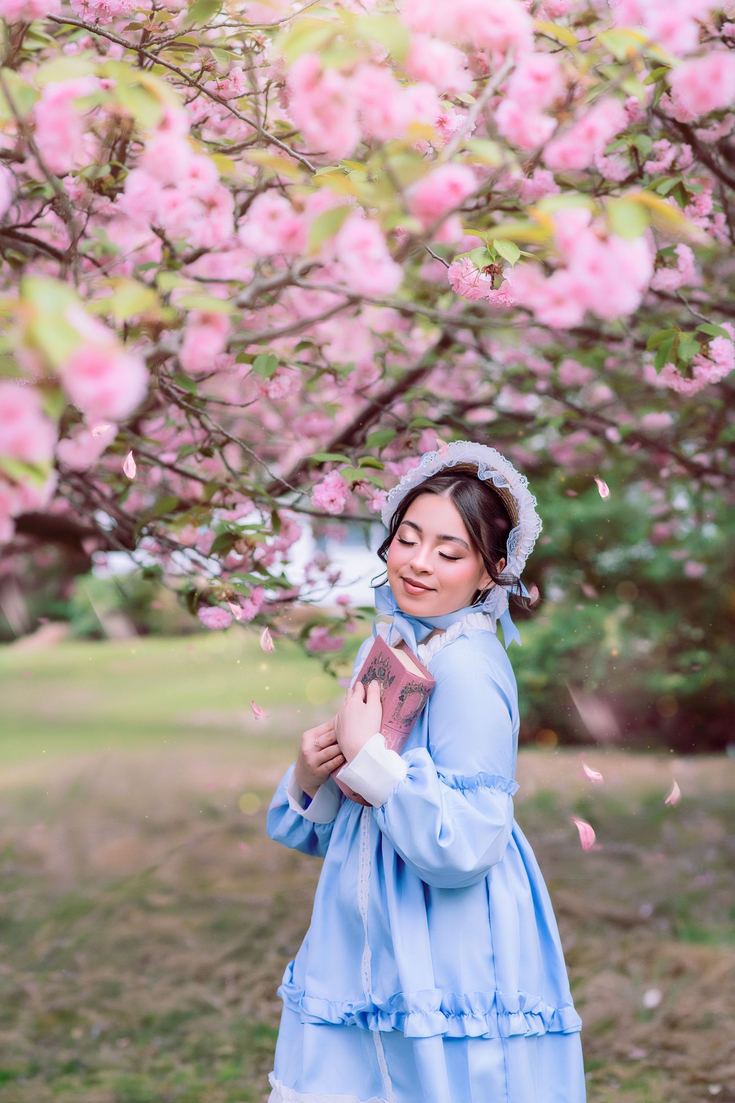 Young woman in blue dress and bonnet hugging a book under blooming pink cherry blossom trees in spring.
