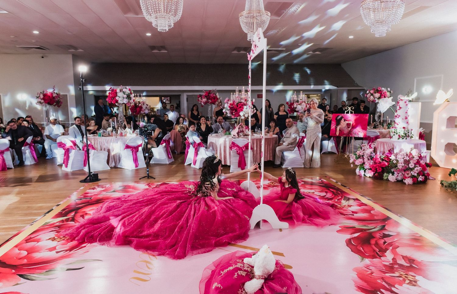 Elegant quinceañera celebration with girls in pink ball gowns dancing on a floral dance floor in a decorated banquet hall.
