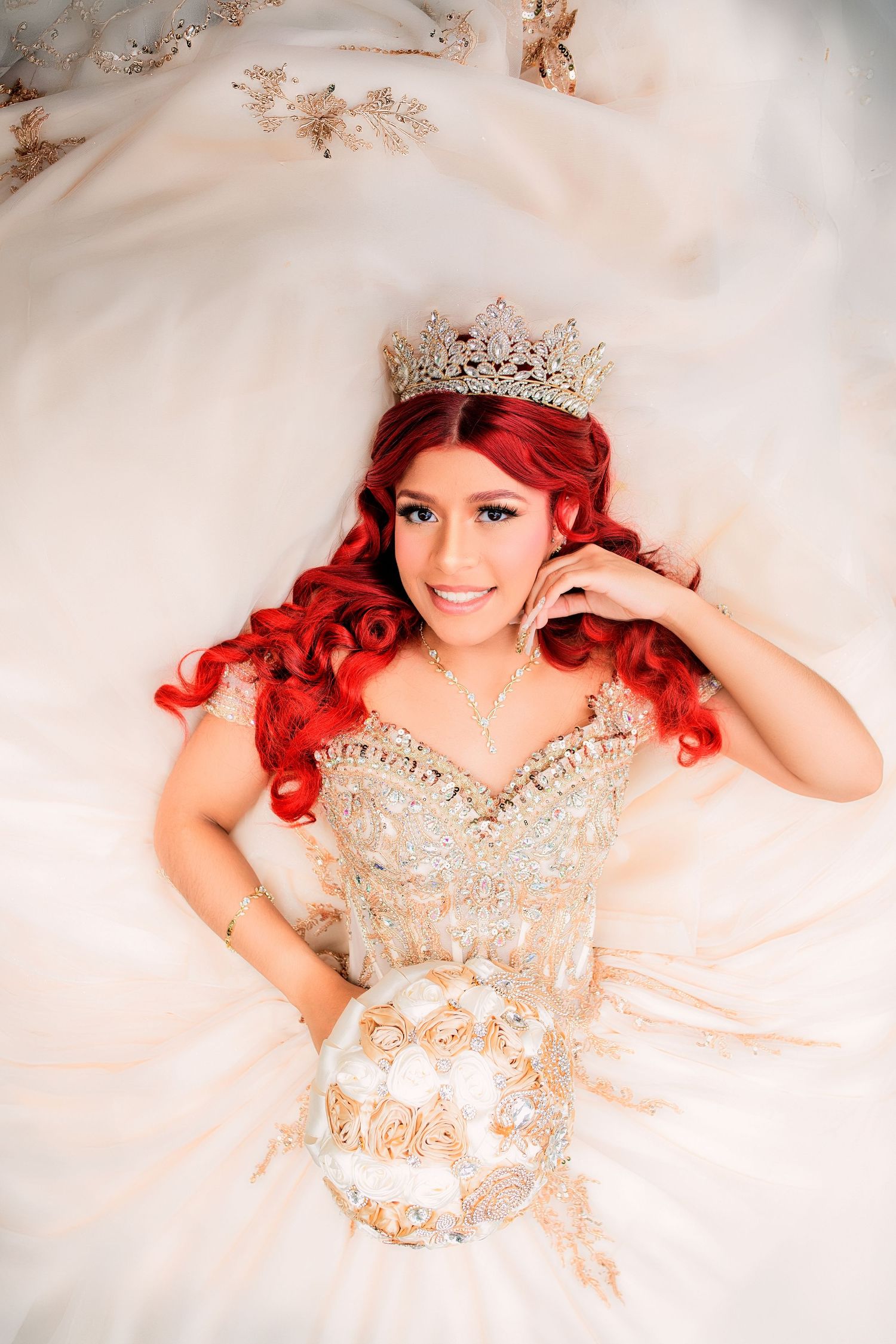 Young woman with red curly hair wearing a crystal crown and ornate white gold quinceañera gown holding a bouquet.