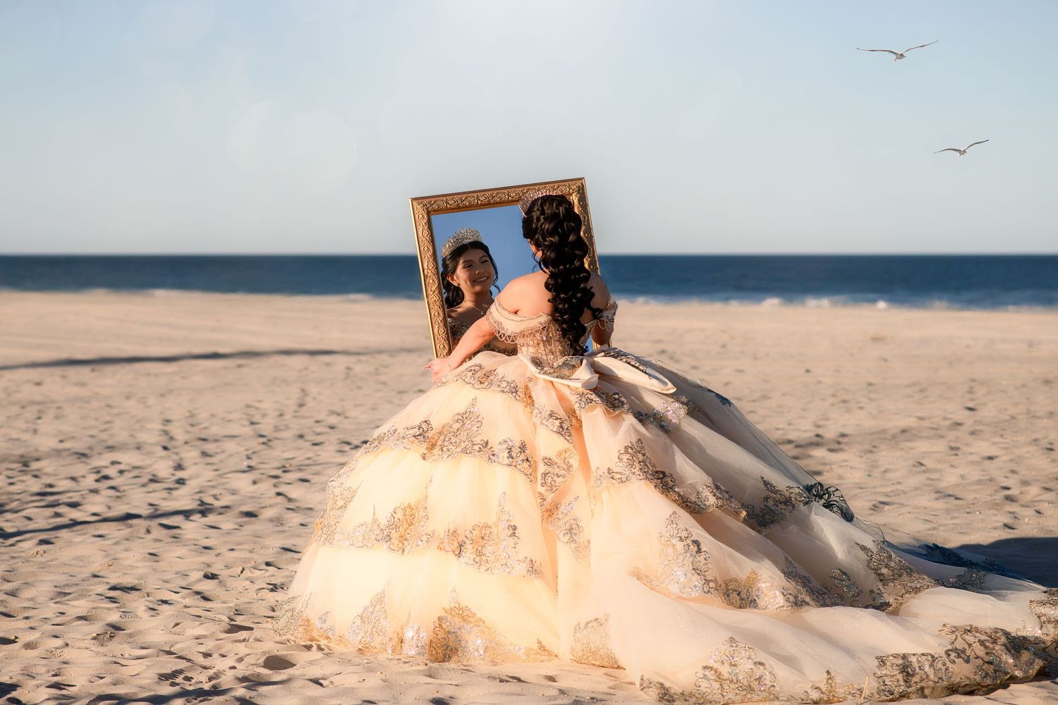Woman in elegant ballgown holding ornate gold frame on sandy beach with ocean backdrop.