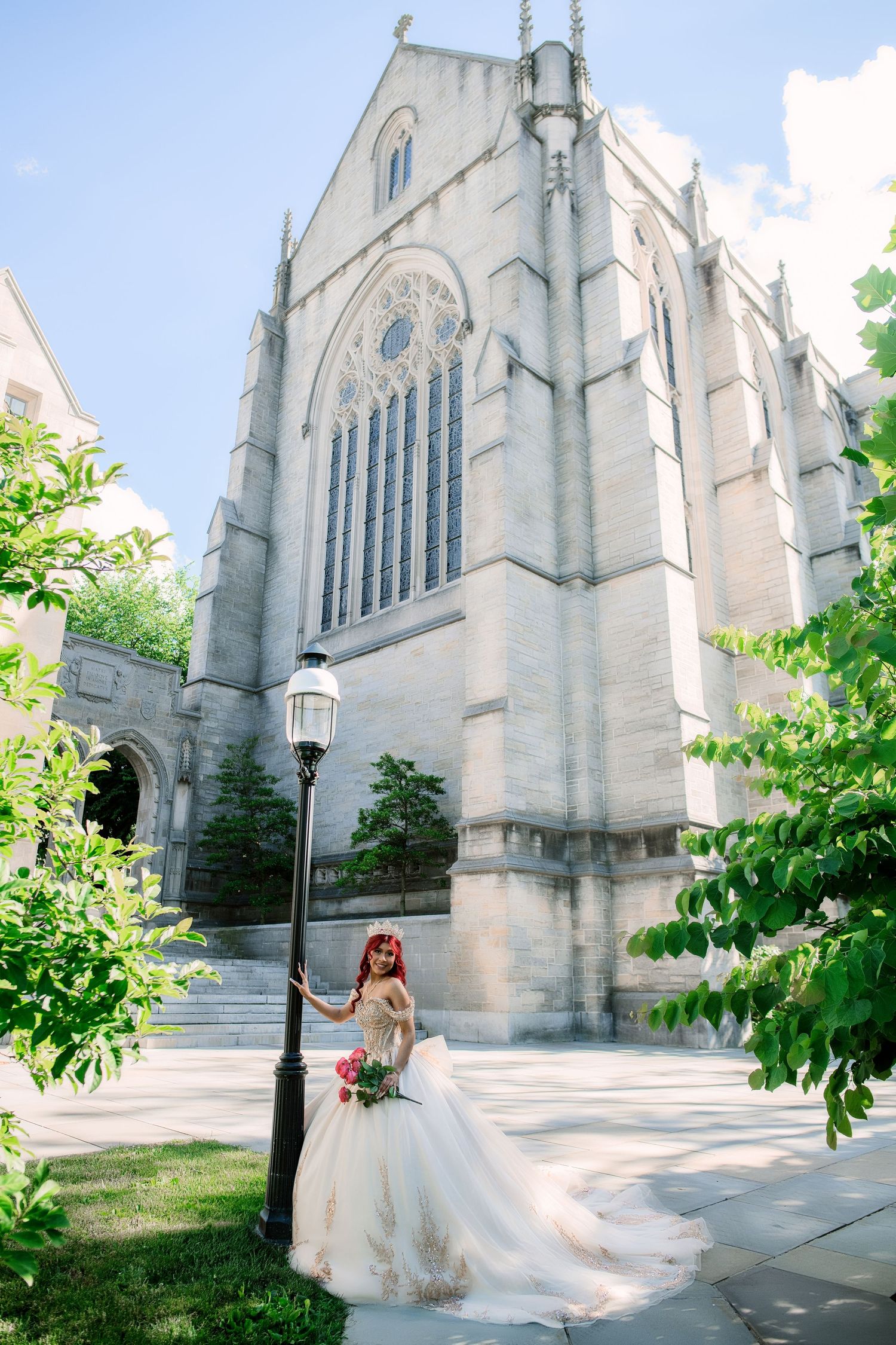 Bride in a flowing white gown poses by a lamp post in front of a grand Gothic stone church on a sunny day.