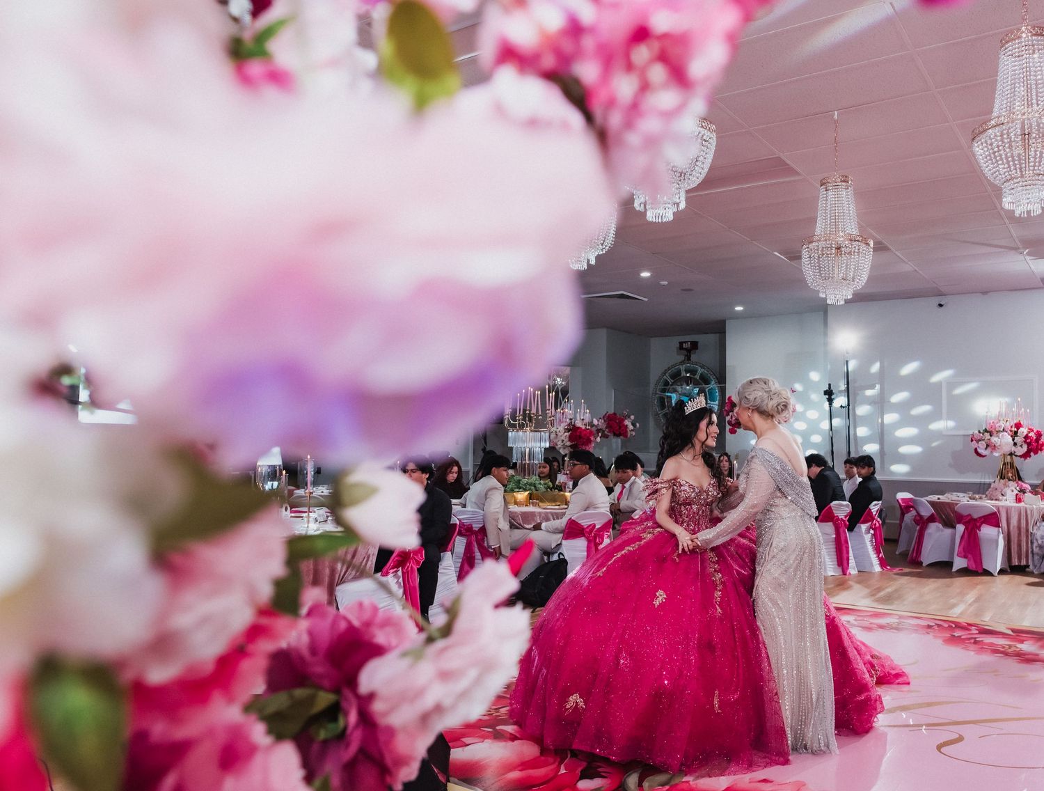 Elegant quinceañera celebration with girl in pink ball gown dancing in decorated ballroom with floral arrangements.