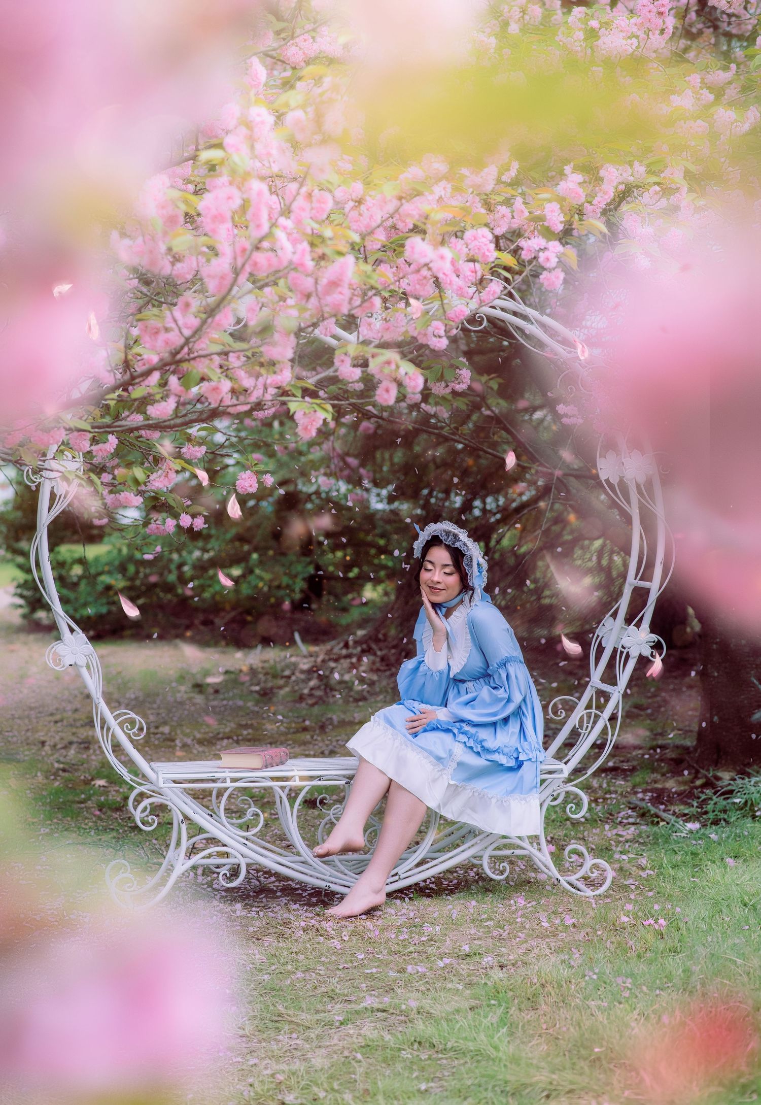 A woman in a blue dress sits on a white ornate bench surrounded by blooming pink cherry blossom trees.
