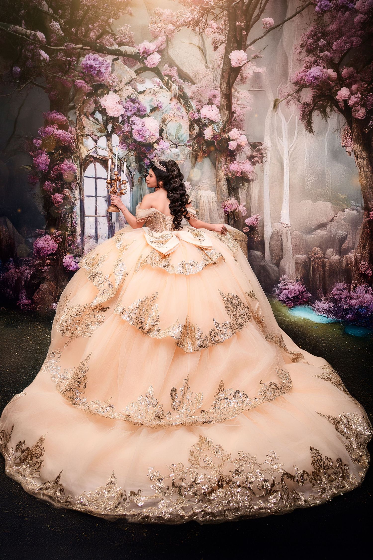 A young woman poses in an ornate champagne gold quinceañera ball gown with lace trim in an enchanted floral fantasy setting.