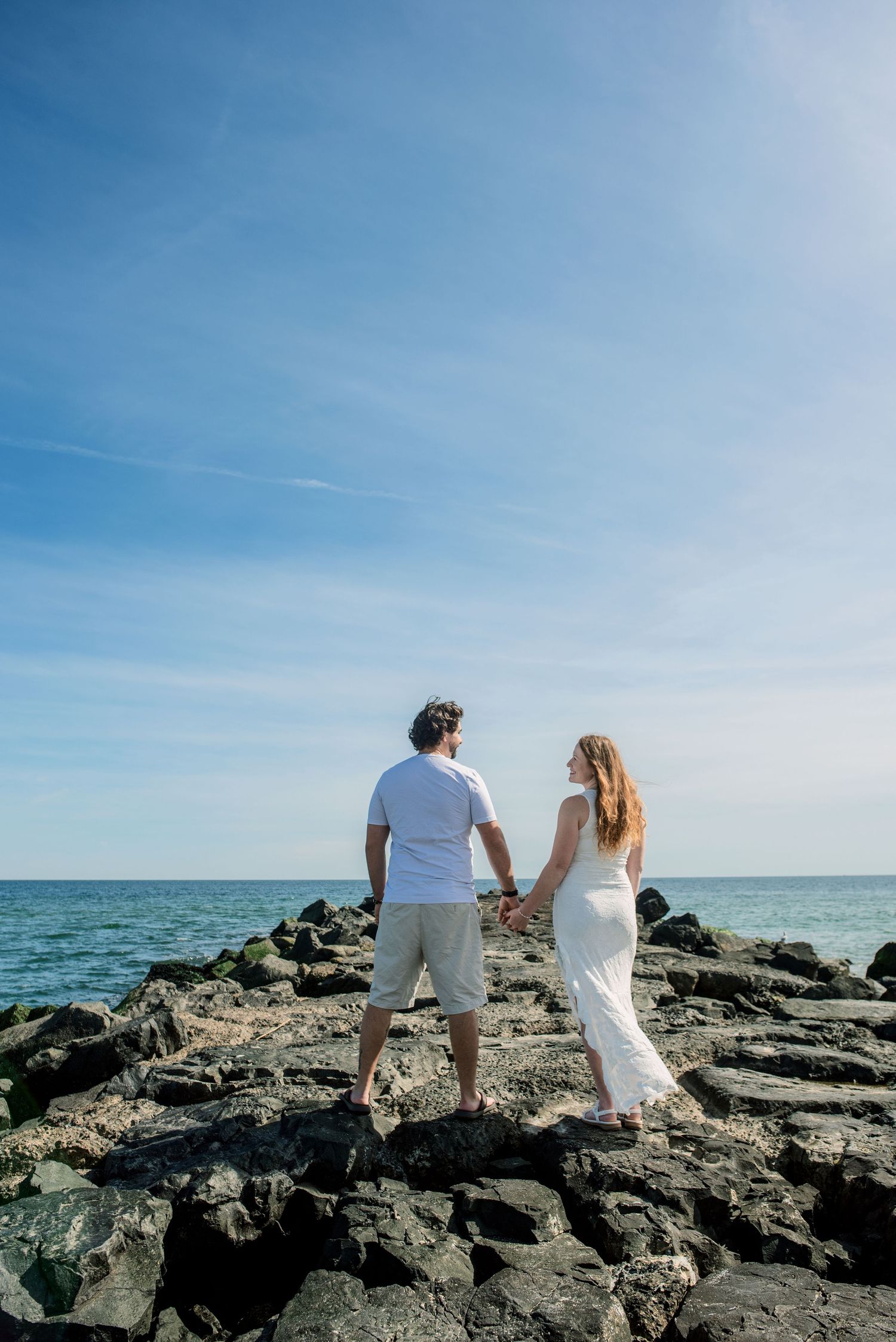 Couple holding hands on rocky shoreline in white outfits, gazing at blue ocean under a clear sky.