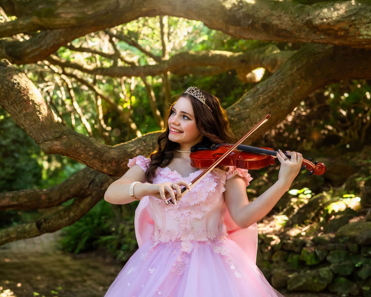 Young woman in pink ball gown playing violin outdoors under scenic tree branches in a enchanting garden setting.