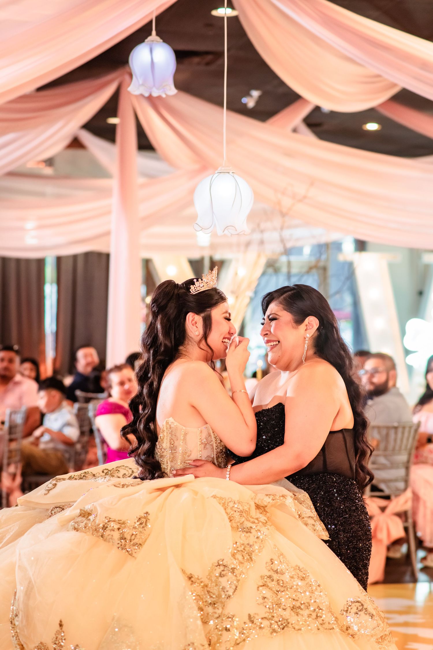 A quinceañera girl in a gold gown shares a tender moment with her mother in a decorated ballroom.