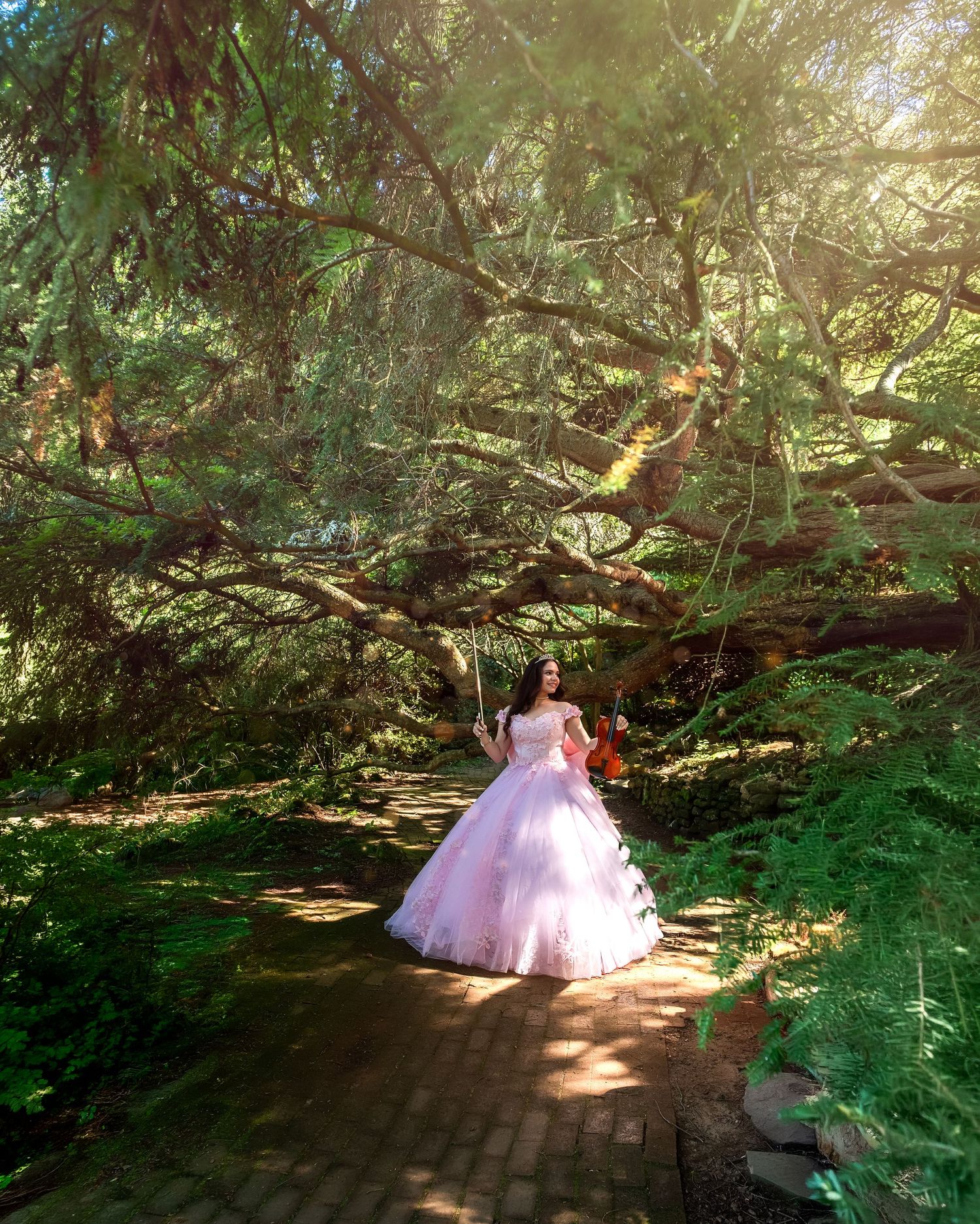 A woman in a pink ball gown holds a violin on a brick path surrounded by lush green trees in a garden.