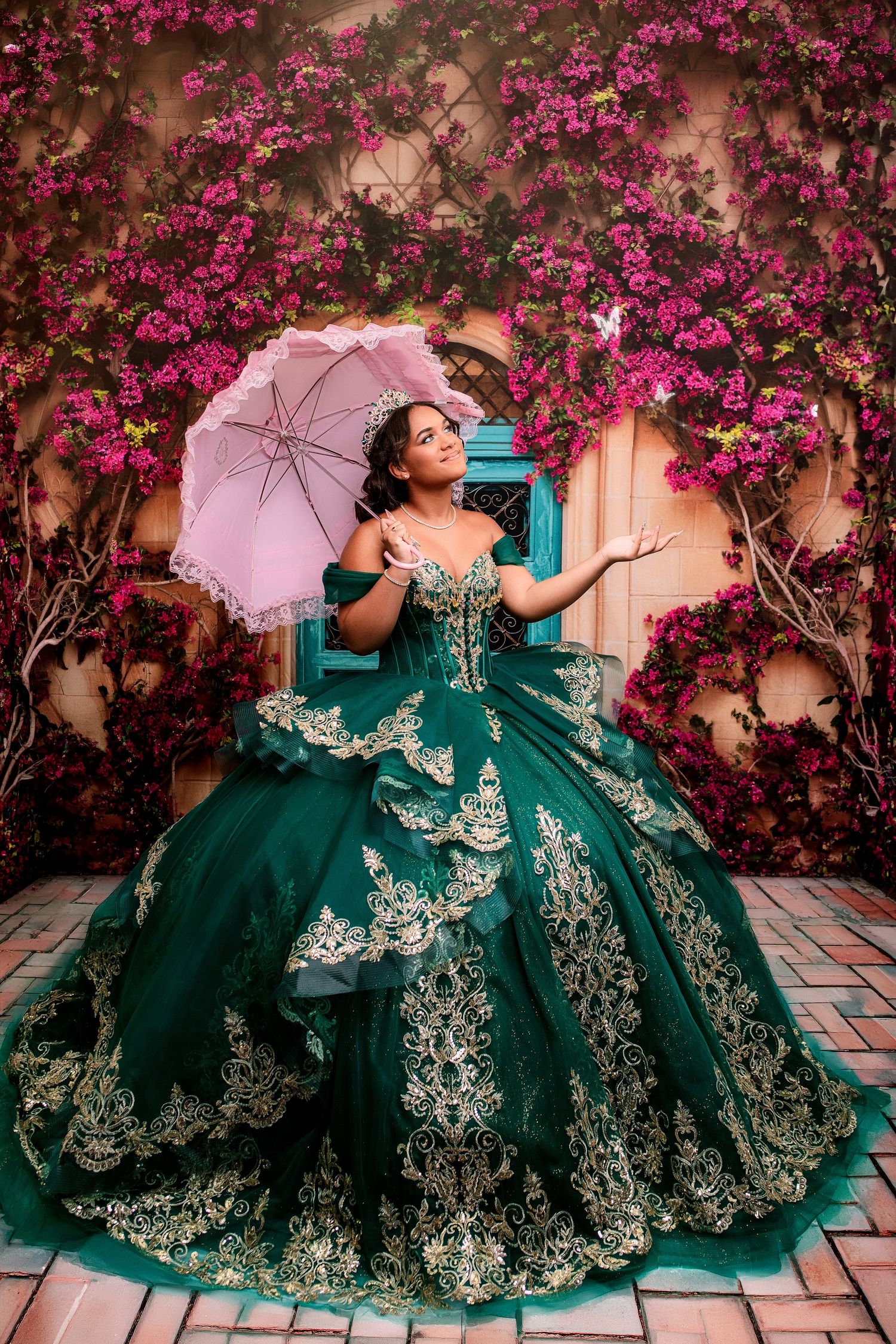 Young woman in emerald green quinceañera ball gown with gold lace, holding pink umbrella amid pink bougainvillea flowers.