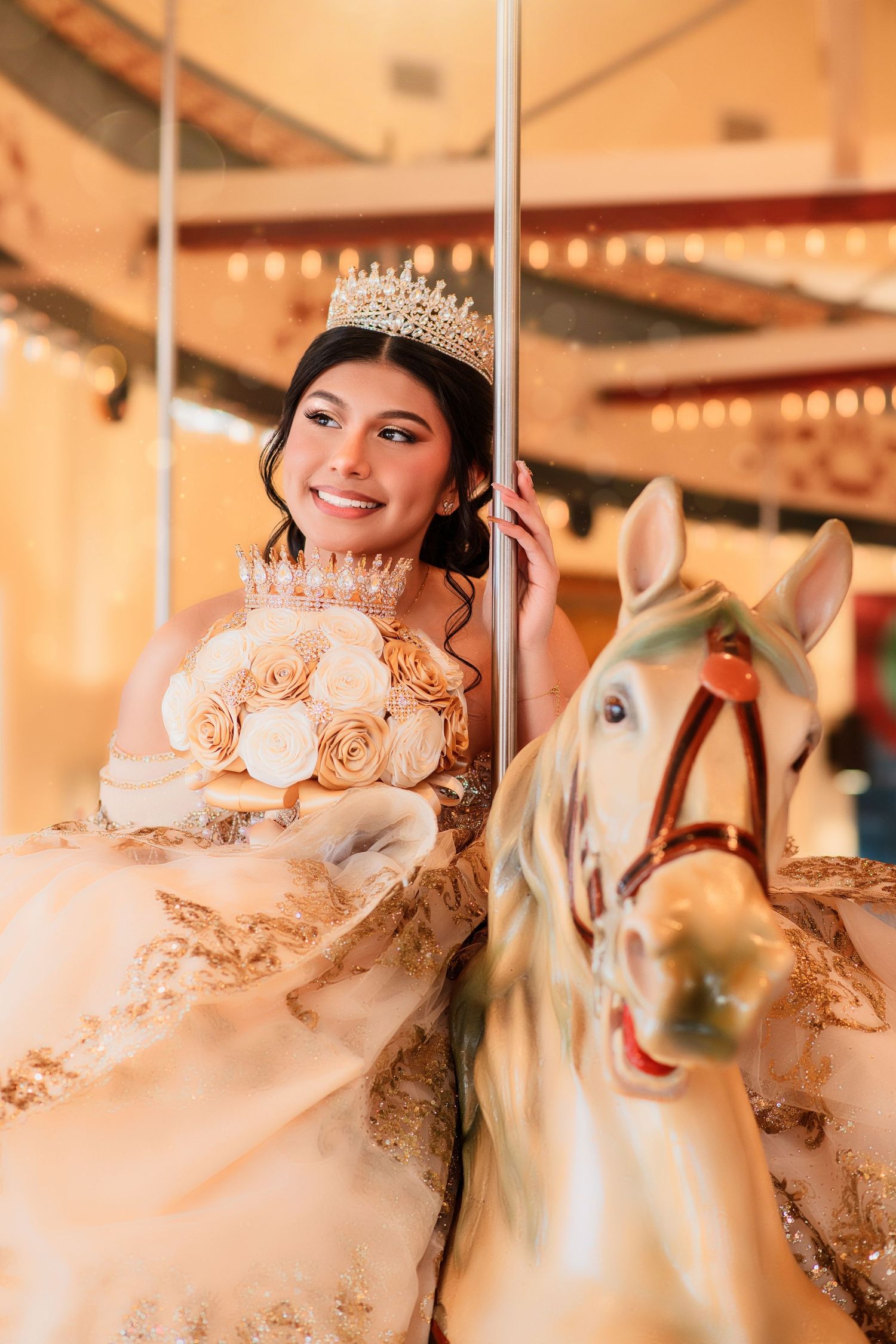 Young woman in gold quinceañera gown and tiara holds rose bouquet while posing on carousel horse.