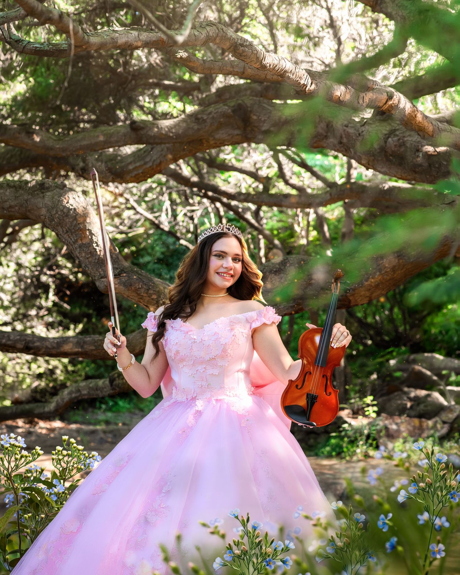 Young woman in pink ball gown holding violin and bow, posing under large tree with blue flowers in enchanted garden.