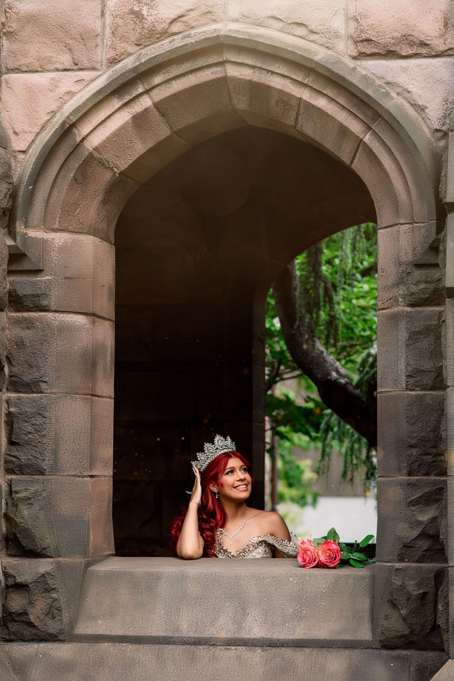 Woman in silver gown and tiara with red hair poses in stone castle arch window with pink roses.