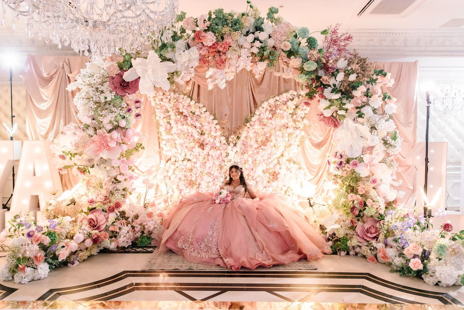 Young girl in pink ball gown sits before giant butterfly floral installation at quinceañera reception.
