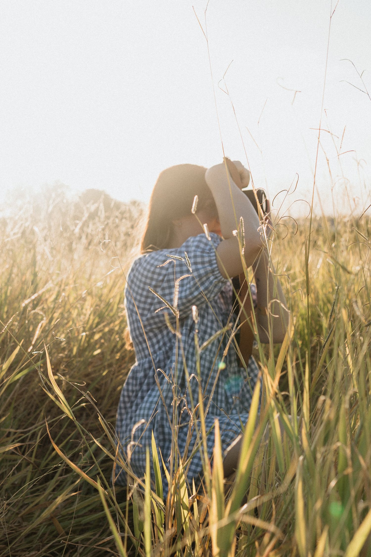 Person in a blue gingham dress stands among tall grass in a golden field during sunset.