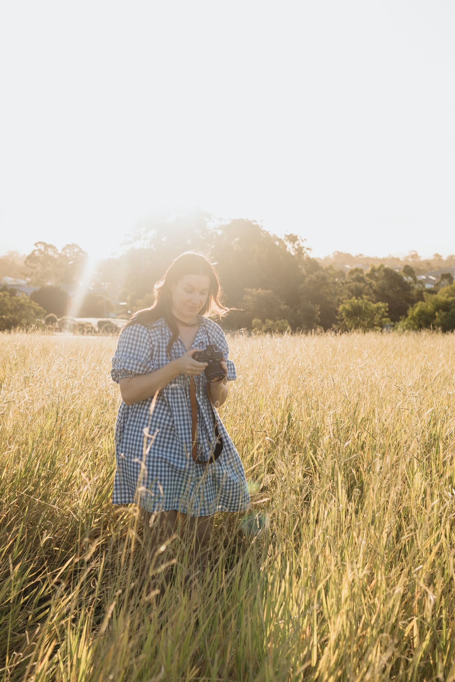 Woman in a blue gingham dress walking through a field and capturing photos with a camera.